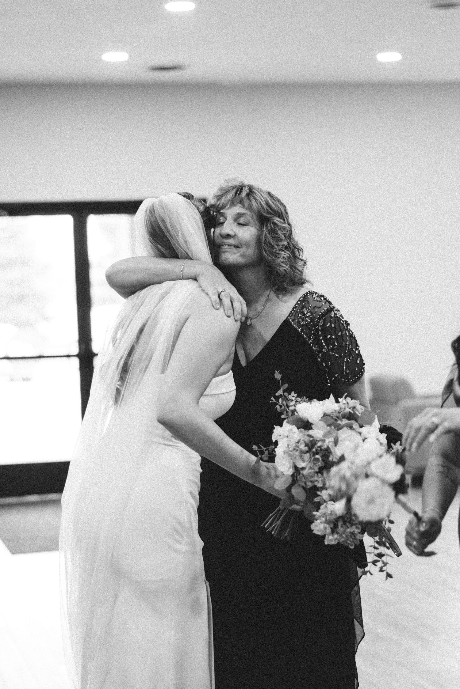 Black and white photo of a bride in a white gown embracing a woman in a dark dress holding a bouquet at a wedding.