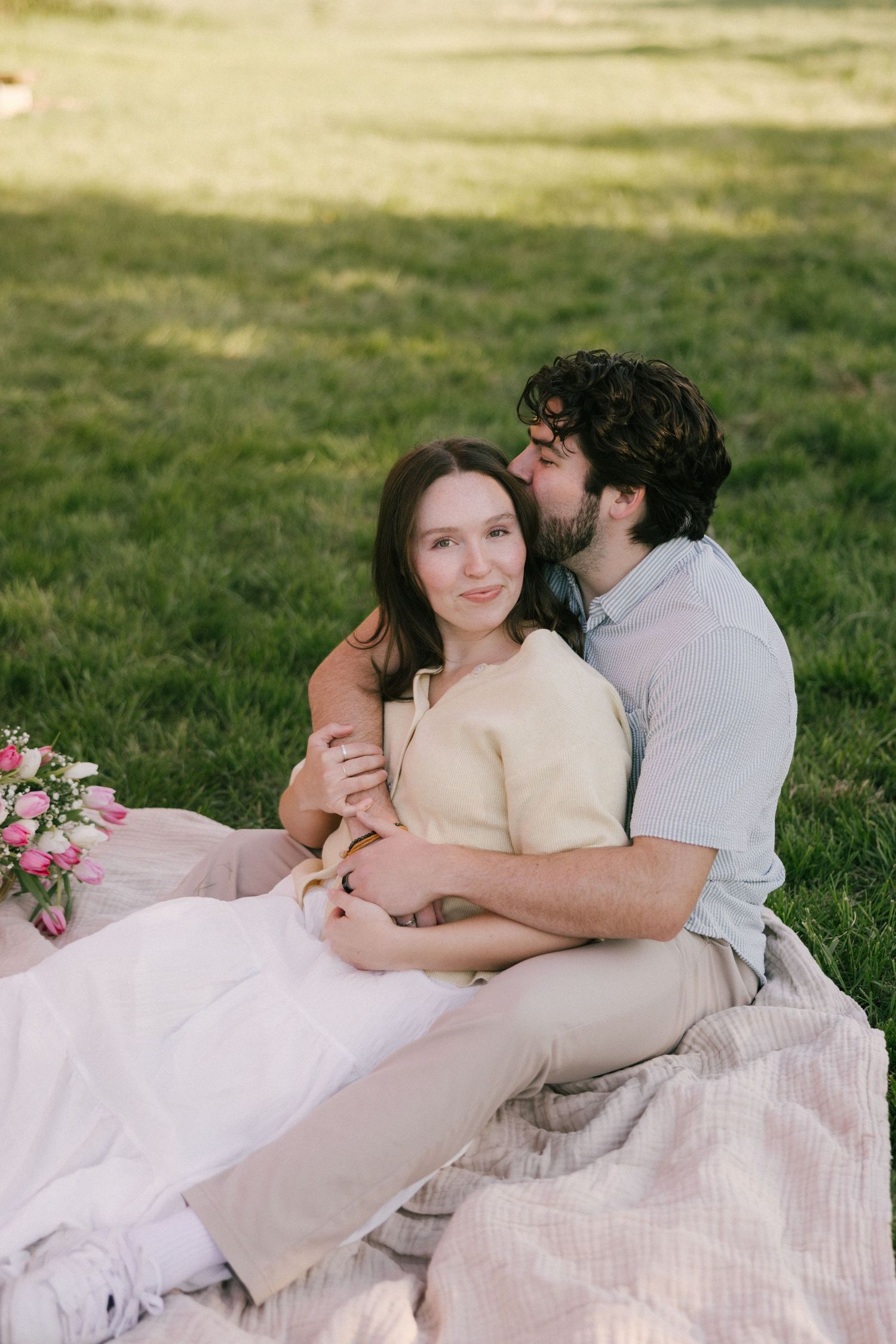 A couple sits on a blanket in a park, the man embracing and kissing a smiling woman with pink flowers nearby.