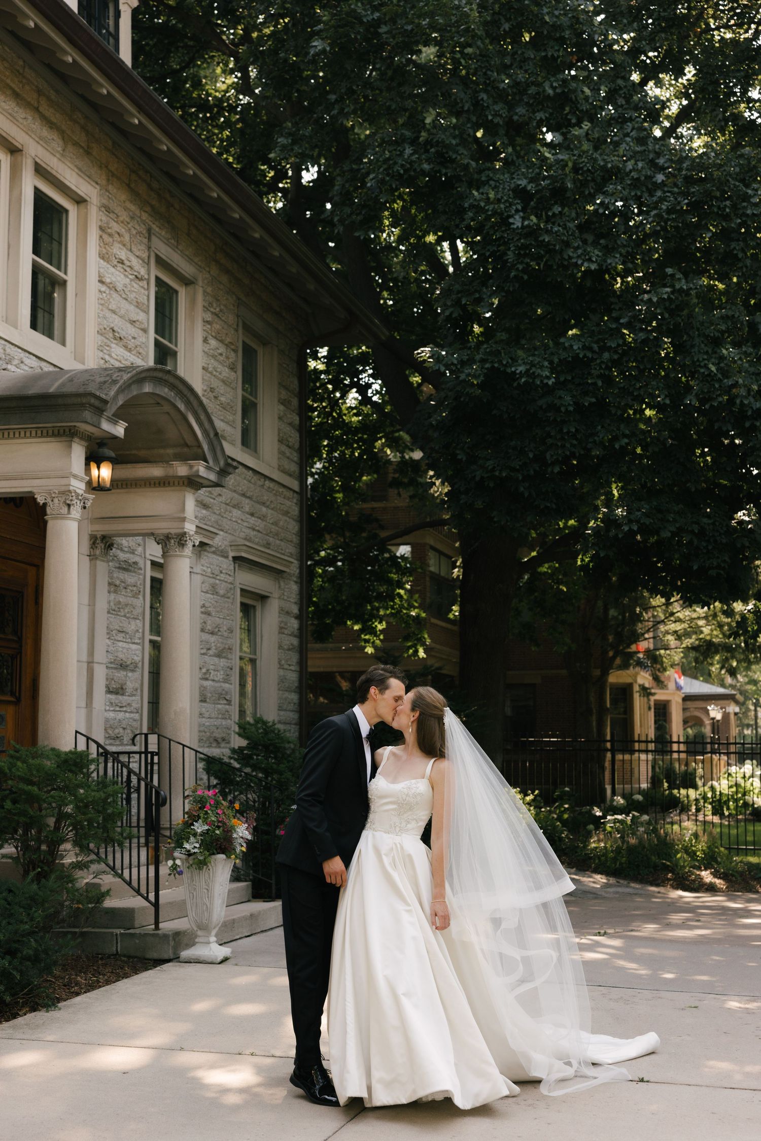 A bride and groom share a kiss outside a stone mansion surrounded by lush trees on their wedding day.