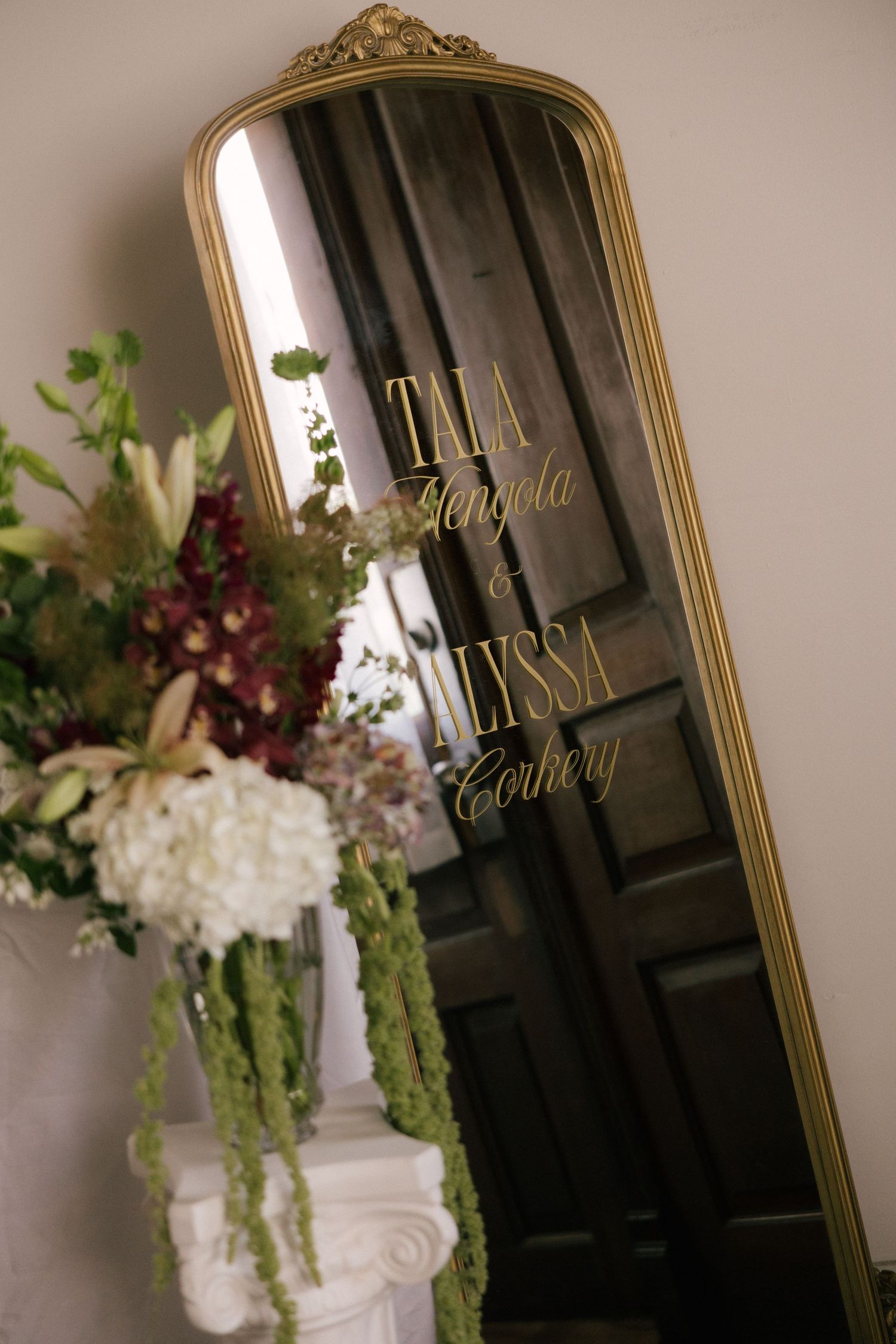 Gold ornate mirror with wedding names Tala Mengola and Alyssa Corkery, beside elegant floral arrangement.