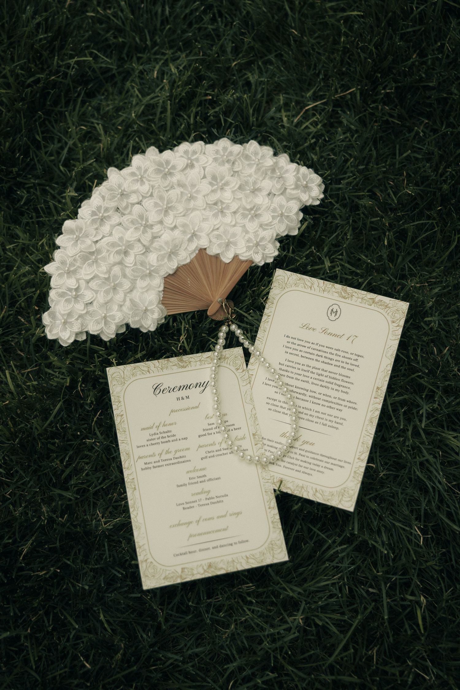 Elegant wedding ceremony programs and a white floral hand fan arranged on lush green grass with a pearl bracelet.