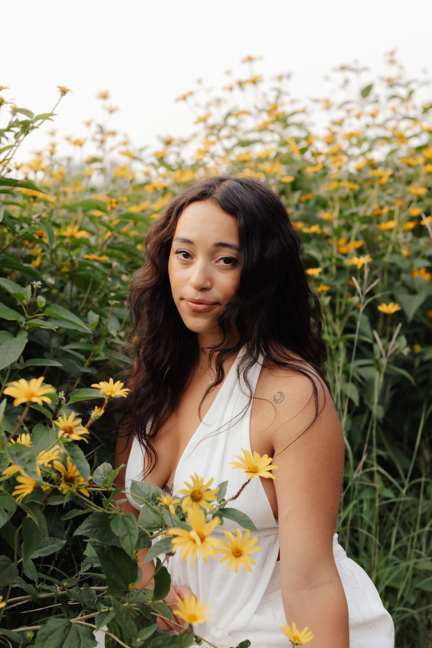 Figure in a white dress poses gracefully among blooming yellow daisies in a summer garden.