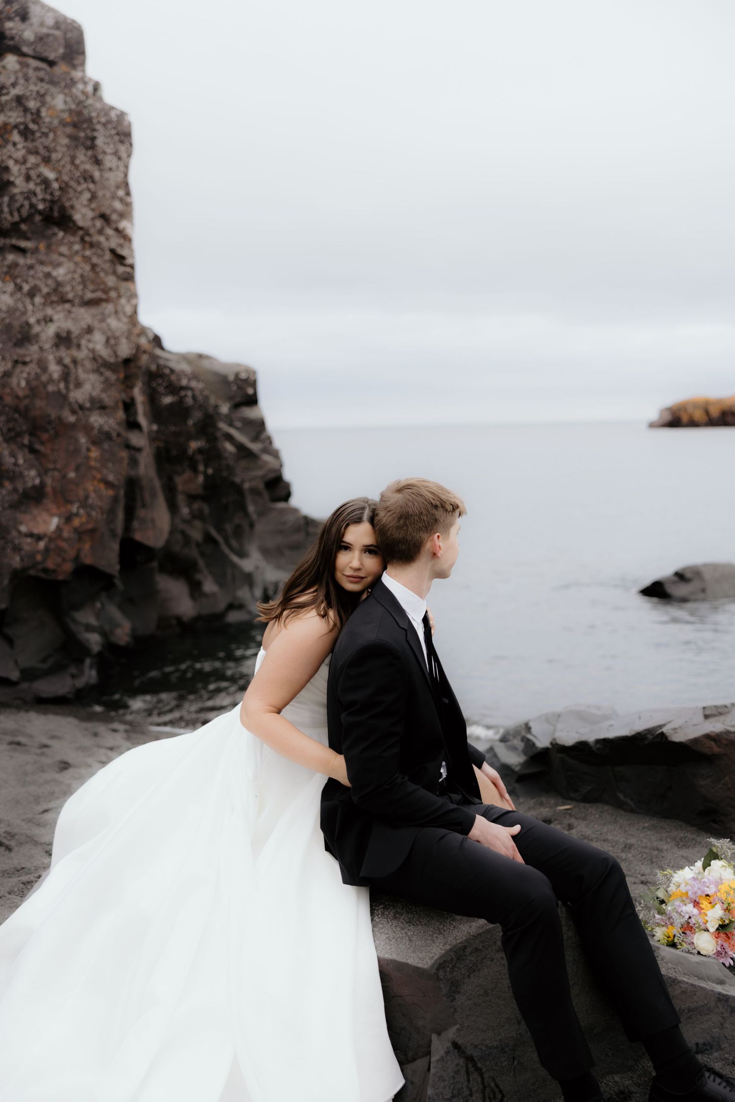 A couple sits together on rocky coastal cliffs overlooking a misty ocean with a wedding bouquet beside them.