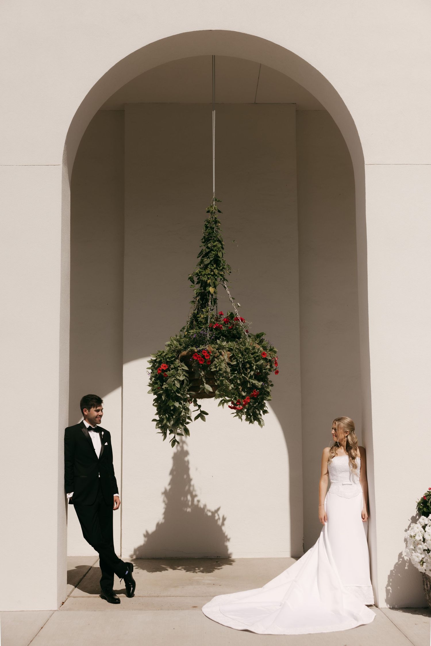 Bride and groom stand beneath a hanging floral arrangement in a white arched alcove during their wedding portrait session.