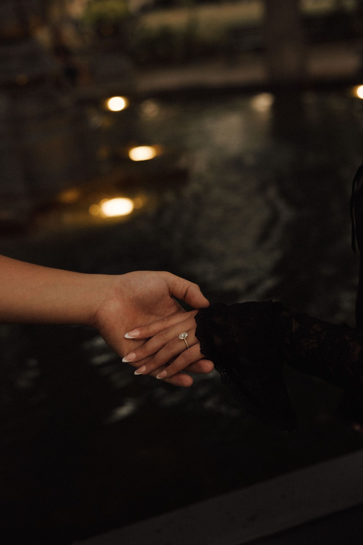 Two hands holding near a dark pool with glowing lights, one adorned with a diamond engagement ring.