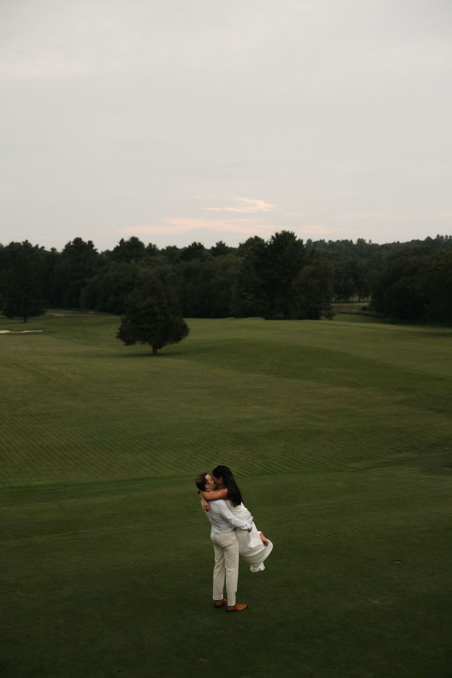 Couple in white embracing on lush green golf course at dusk with trees in background.