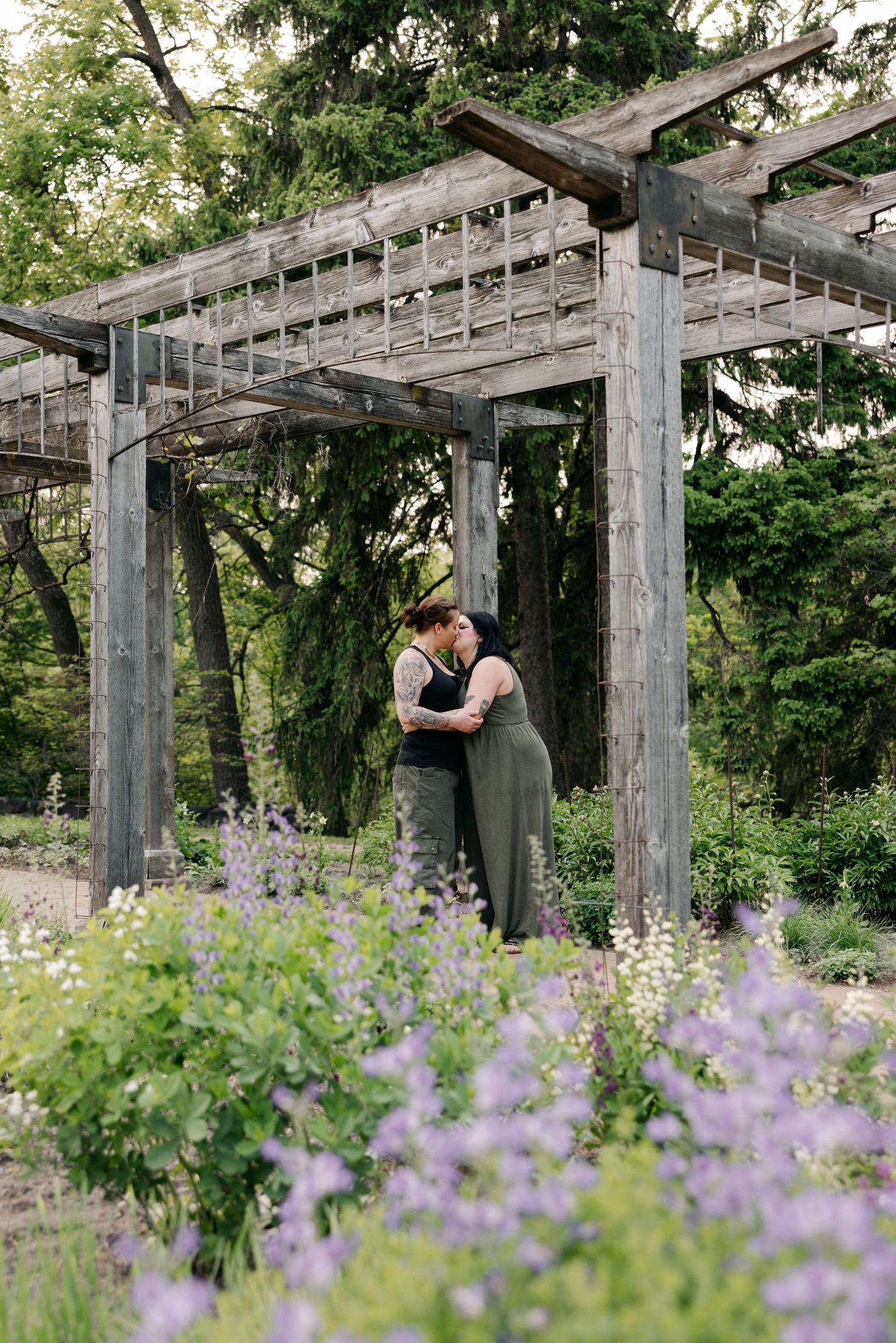A romantic couple shares a tender moment under a rustic wooden pergola surrounded by blooming purple wildflowers in a garden.