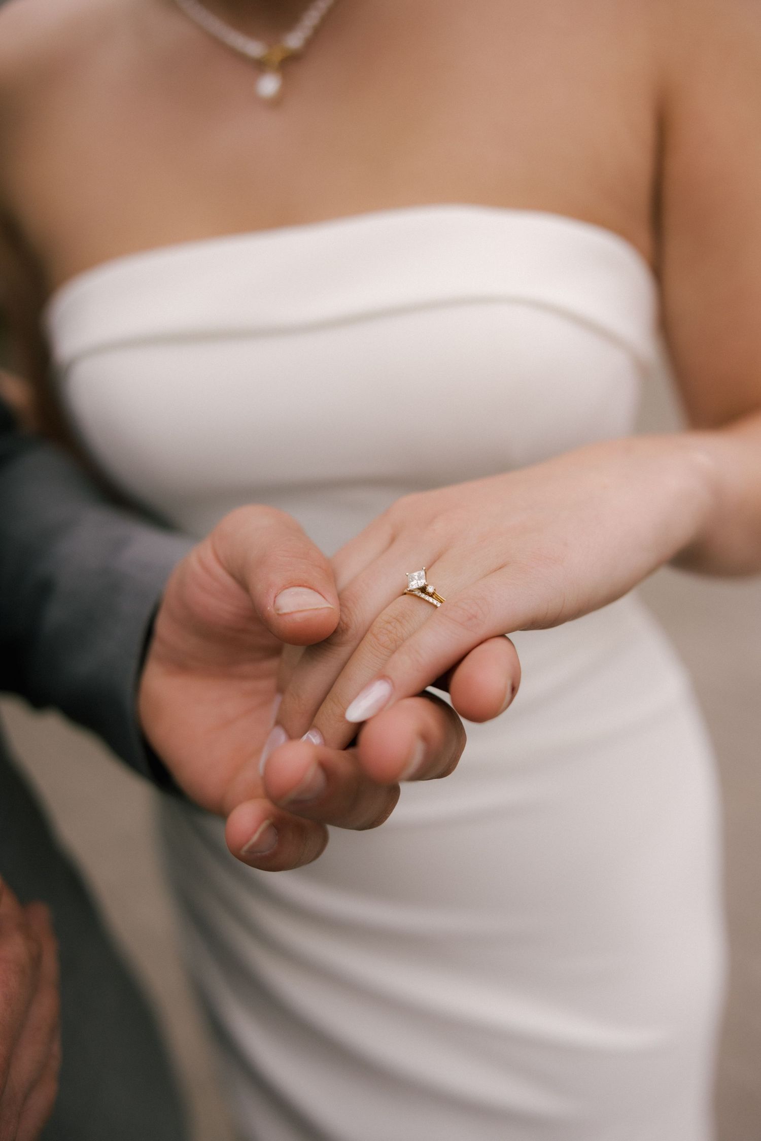 Bride in white strapless gown holding groom's hand, showcasing a delicate gold diamond engagement ring.