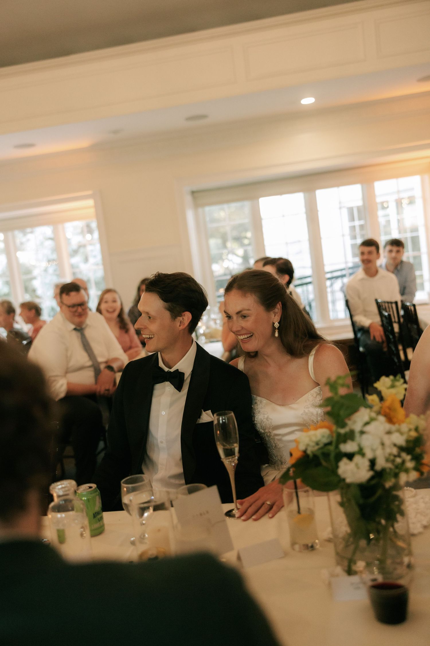 Bride and groom smiling at wedding reception table with champagne flute and floral centerpiece.