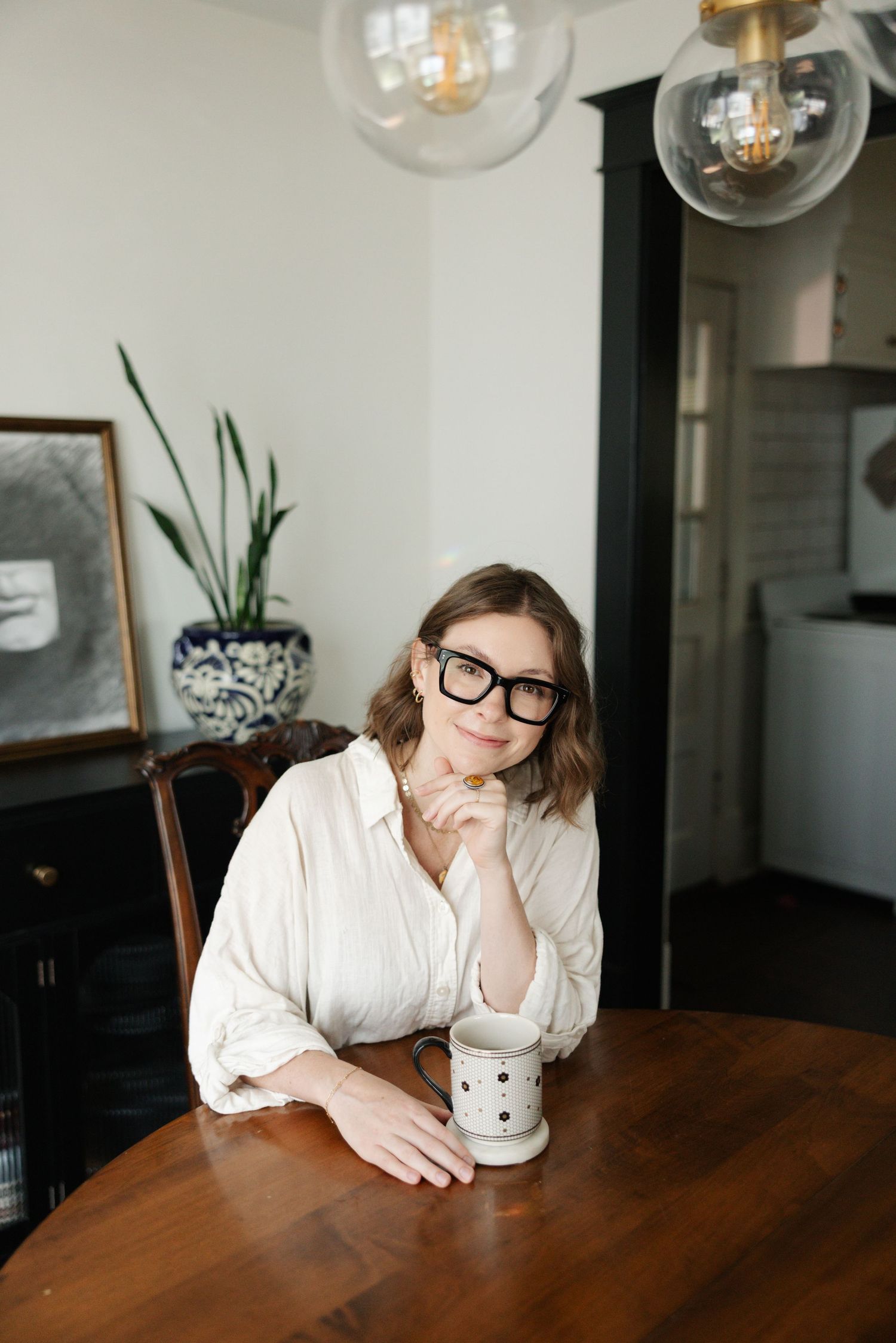 Person in white shirt and glasses rests head on wooden dining table with pendant lights hanging above.