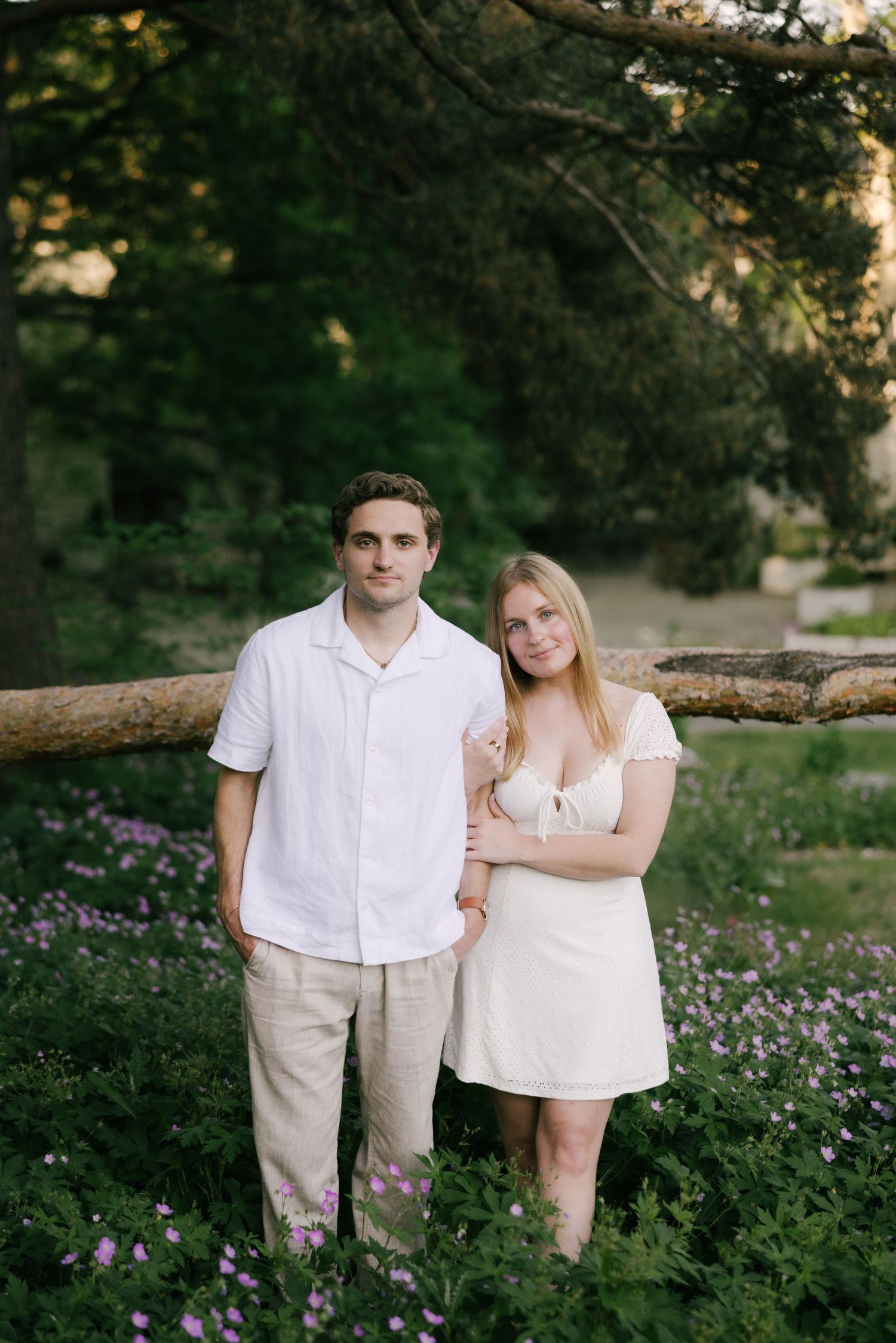 A young couple in white outfits poses among purple wildflowers and lush greenery in a romantic outdoor setting.