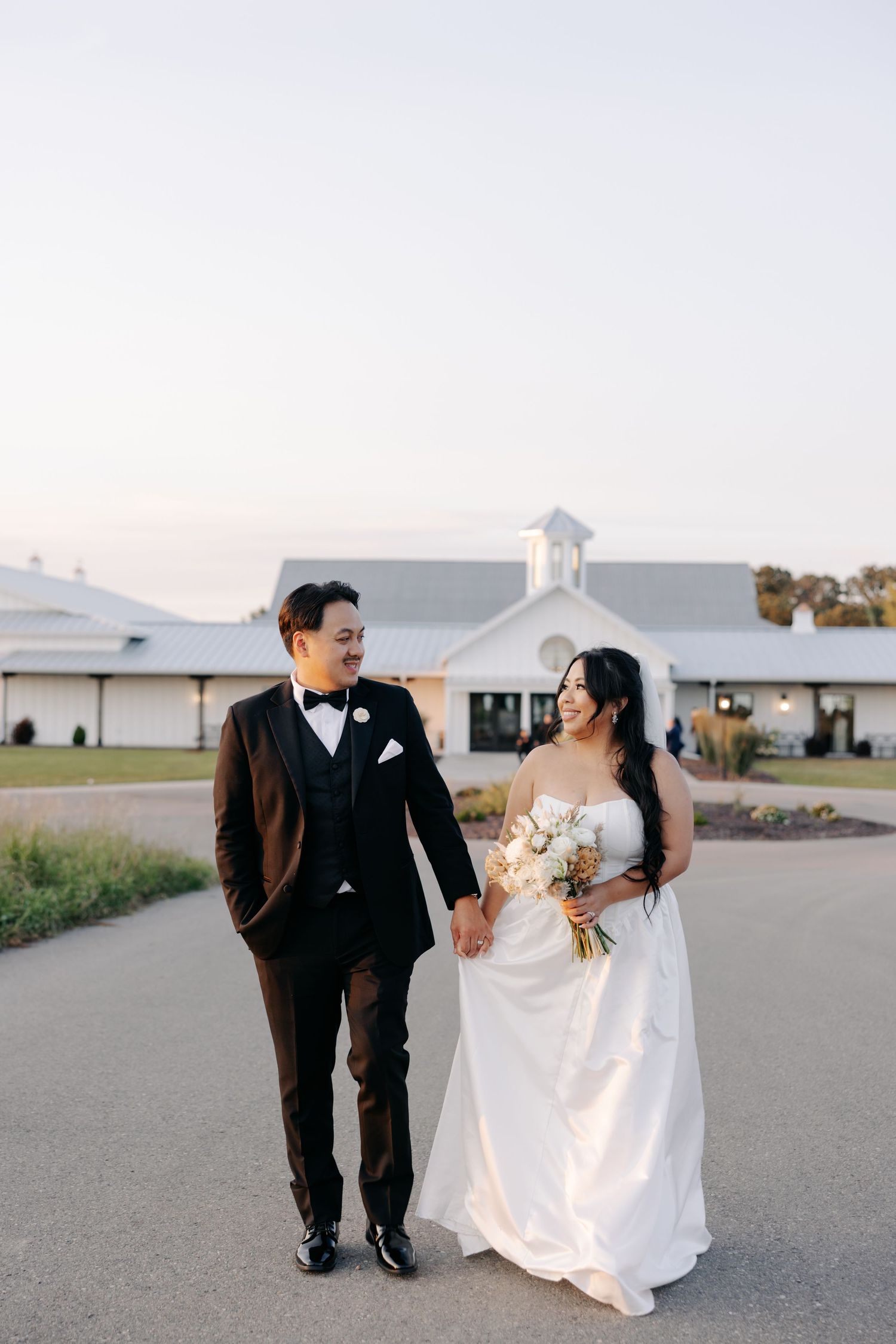 A newlywed couple walks together on a paved road with a white barn-style building in the background during sunset.