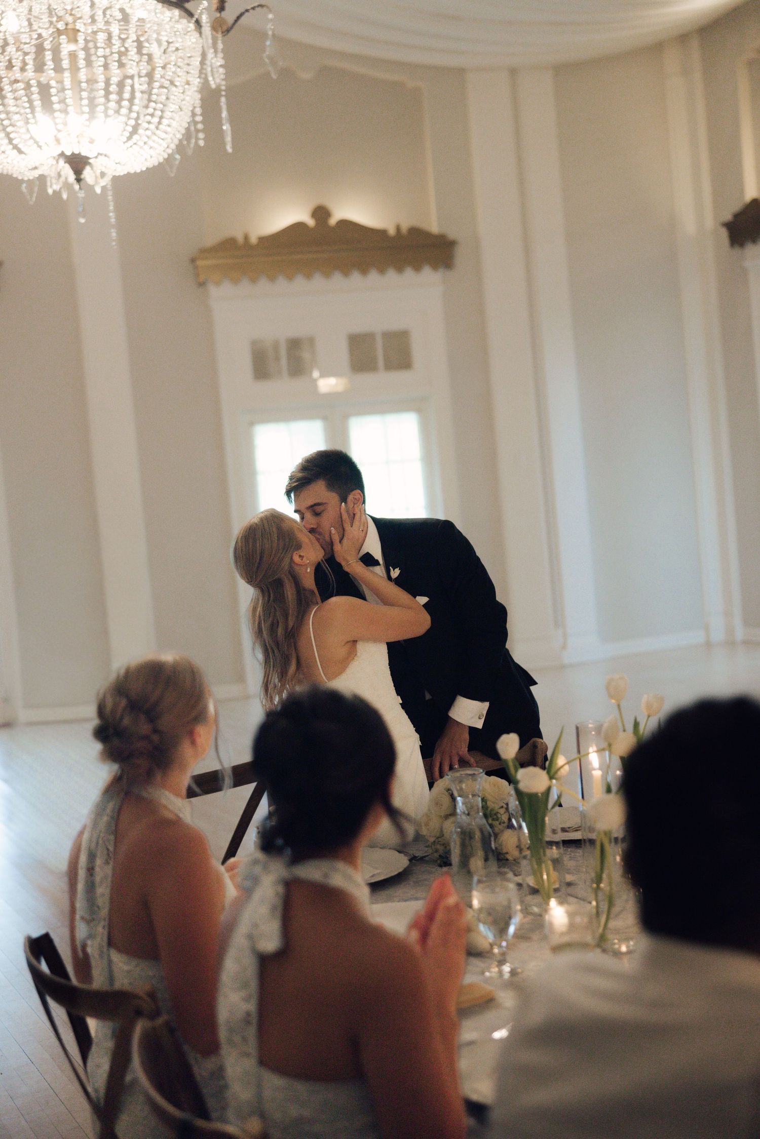 Bride and groom share a kiss at their elegant wedding reception while guests applaud at a beautifully decorated table.