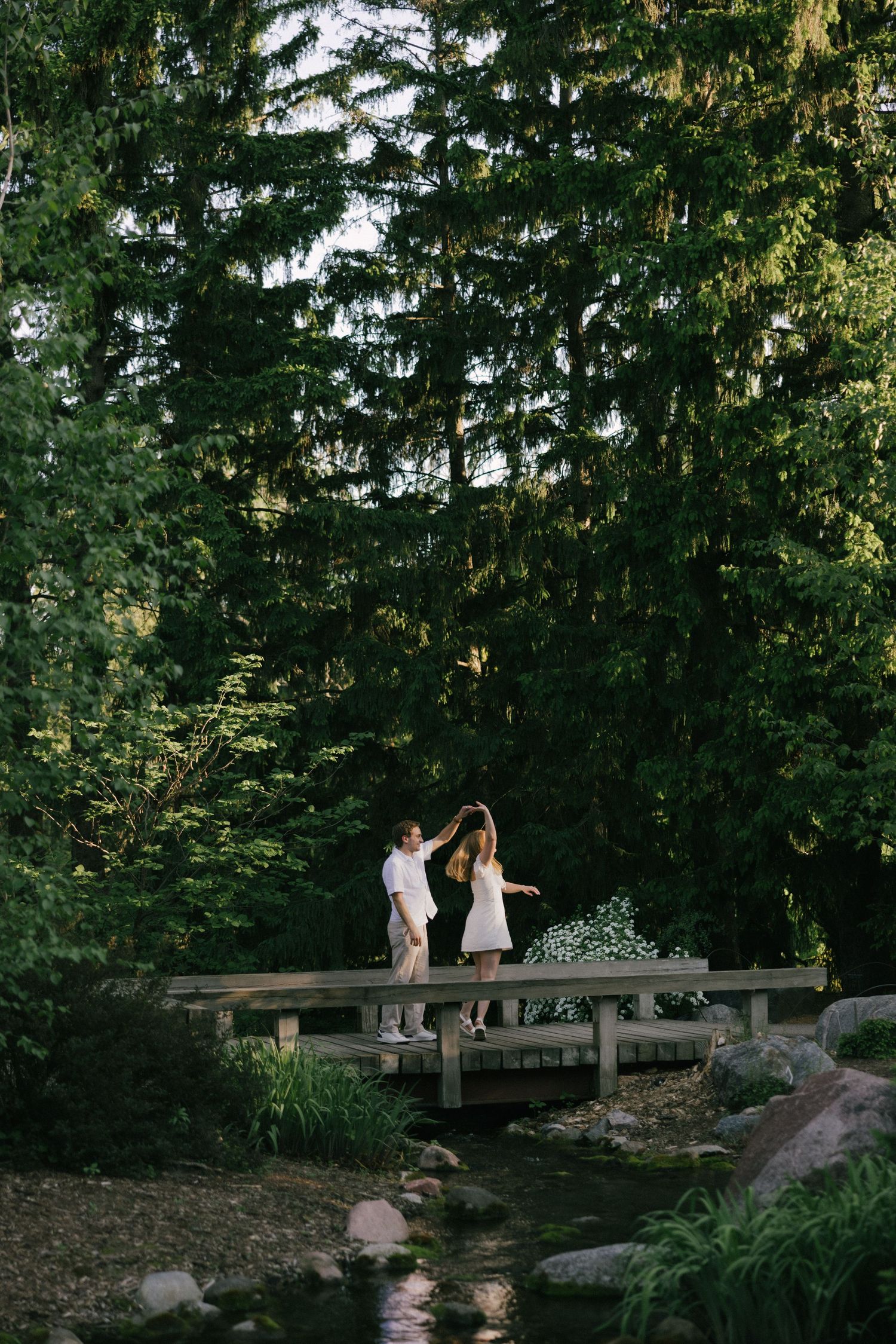 Couple dancing on a wooden bridge surrounded by lush green trees and a flowing stream in a forest setting.