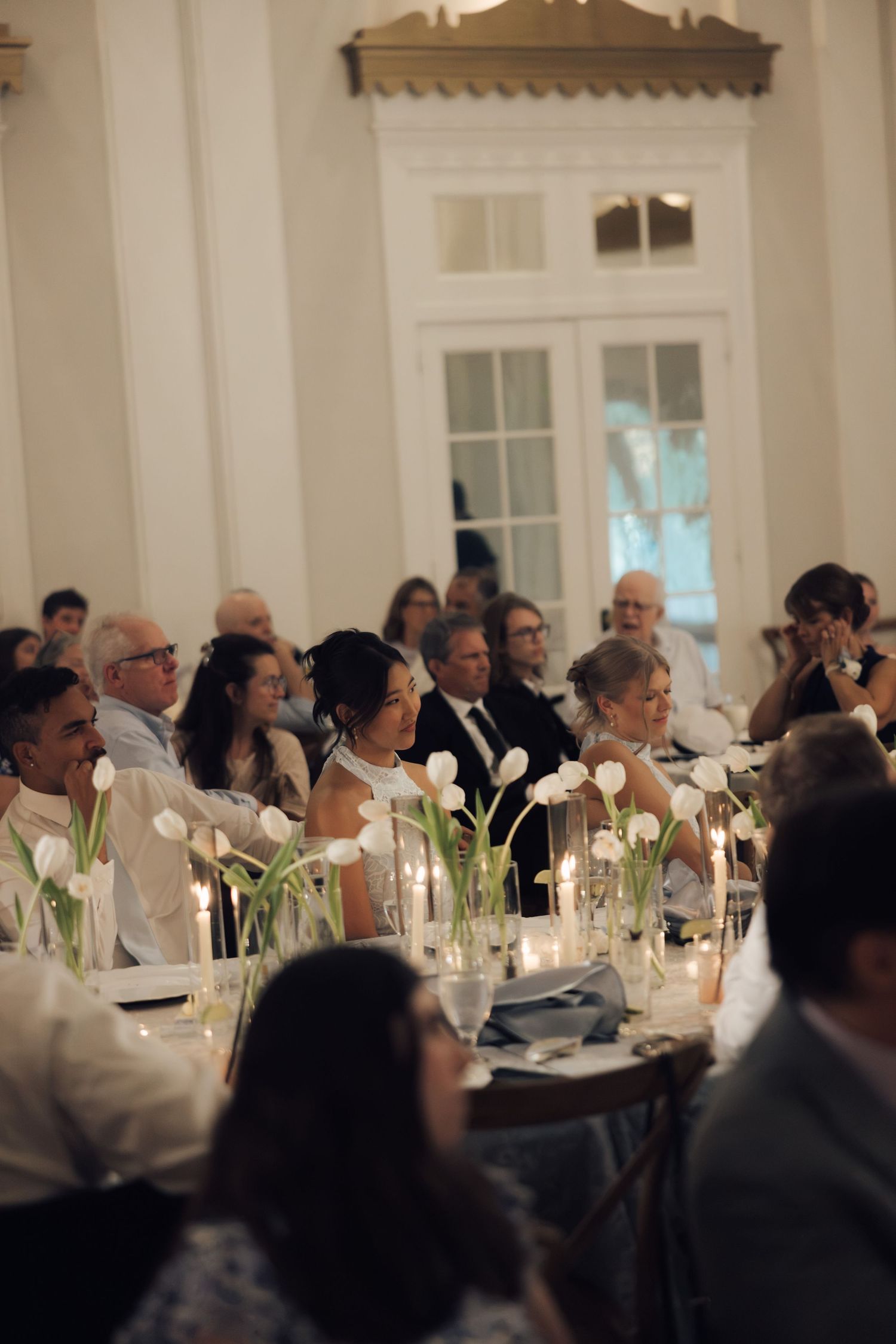 Elegant wedding reception with guests seated at candlelit tables adorned with white tulips in glass vases.