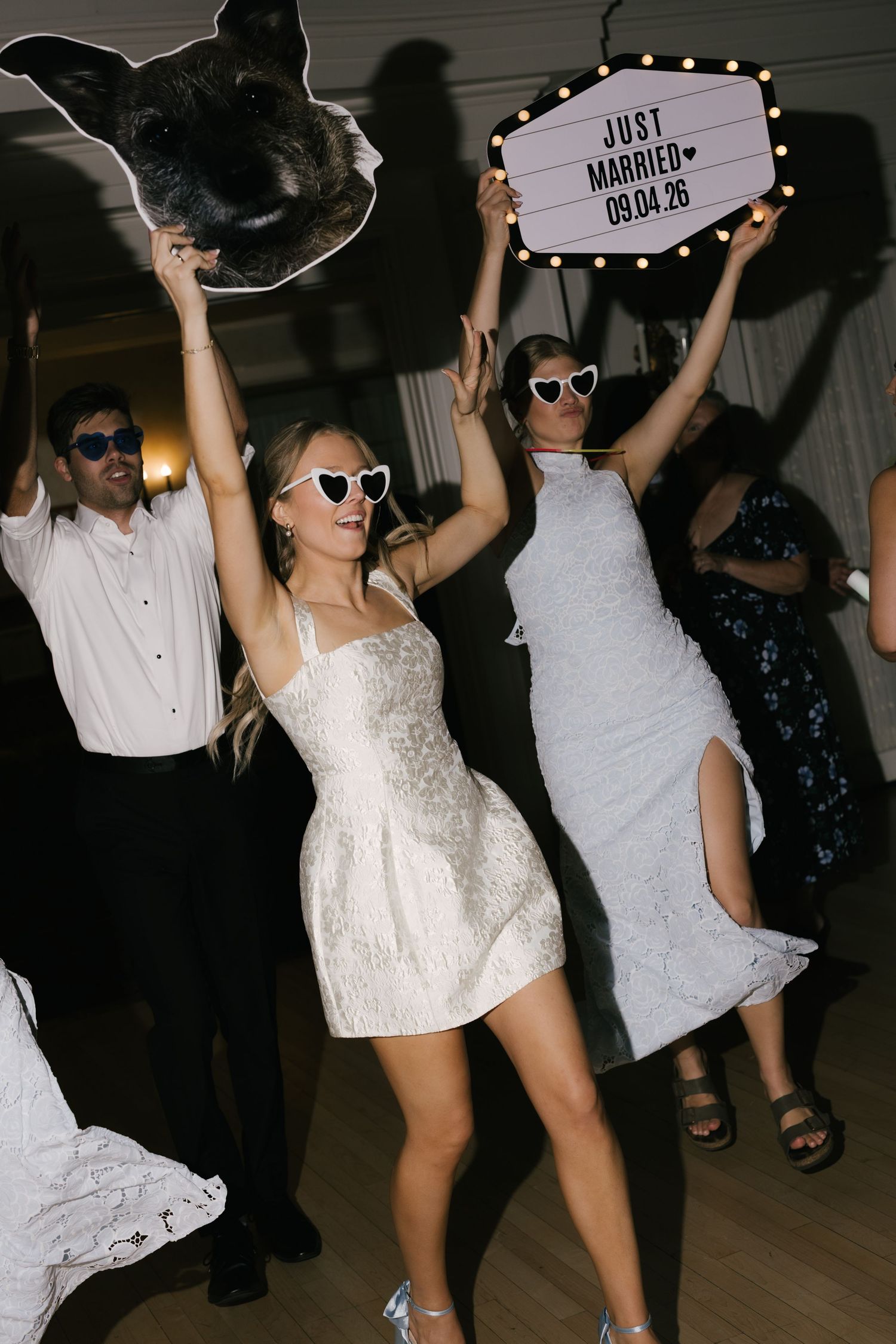 Bride in white mini dress dancing at wedding reception holding dog cutout, Just Married sign dated 09.04.26.