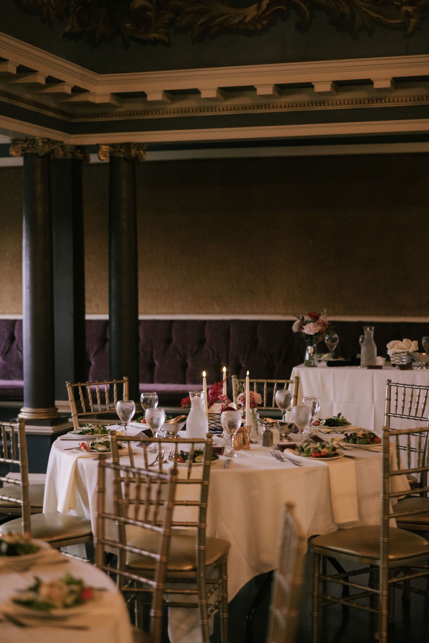 Elegant banquet hall with round tables set for a formal dinner, gold Chiavari chairs, candles, and ornate dark walls.
