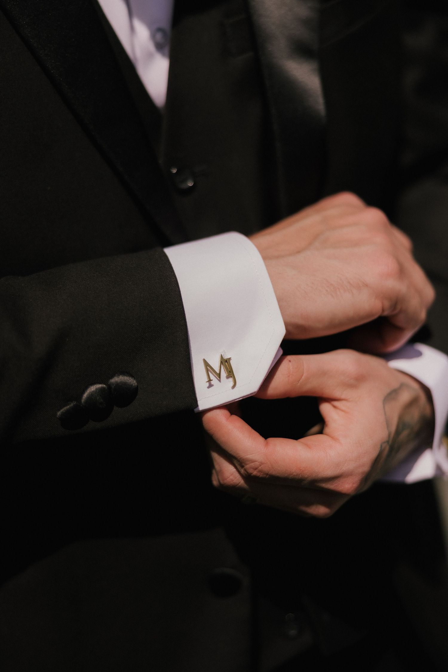 Groom adjusting monogrammed gold MJ cufflinks on white dress shirt with black tuxedo jacket.