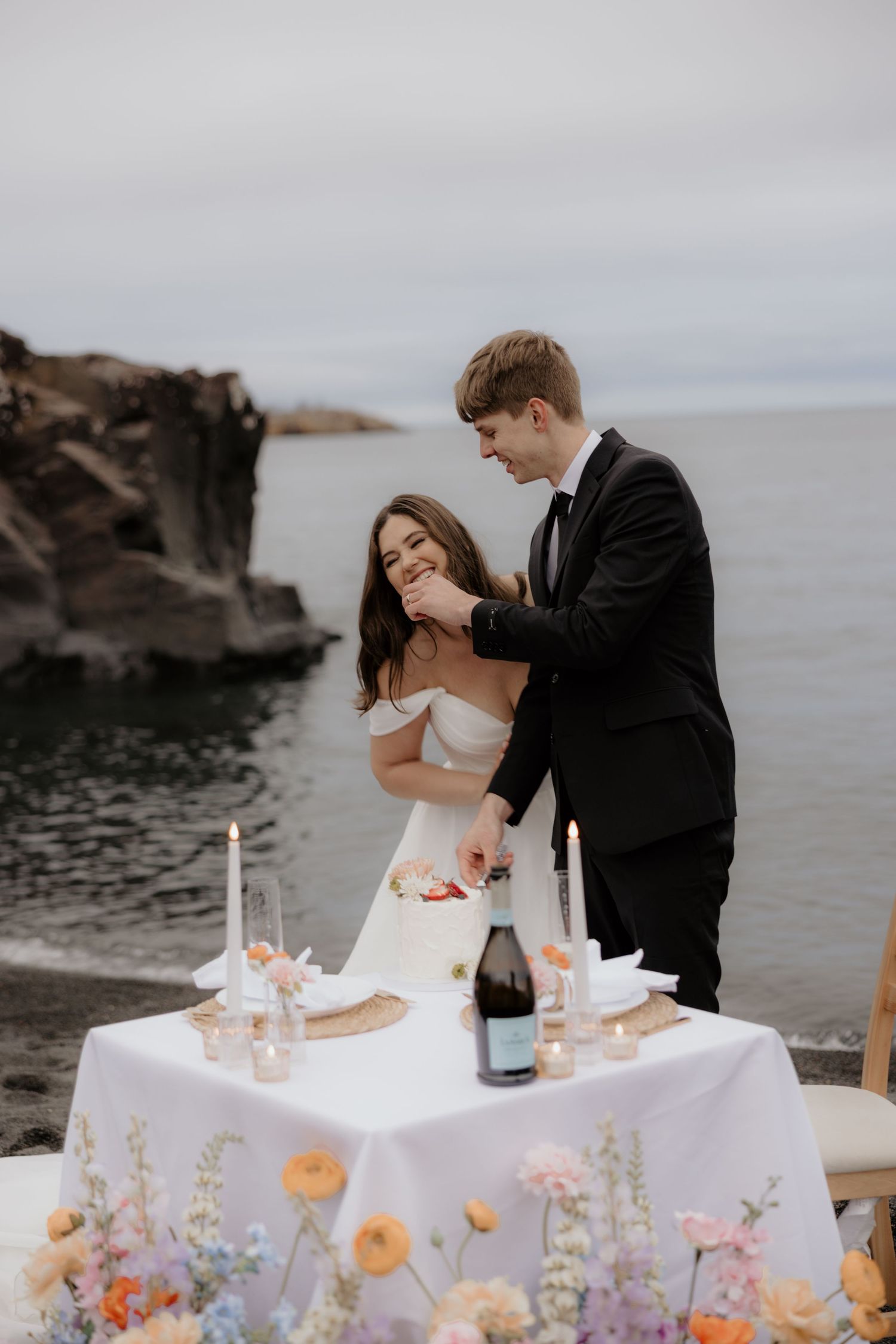 Romantic wedding couple shares intimate moment by candlelit table with champagne on rocky ocean coastline at sunset.