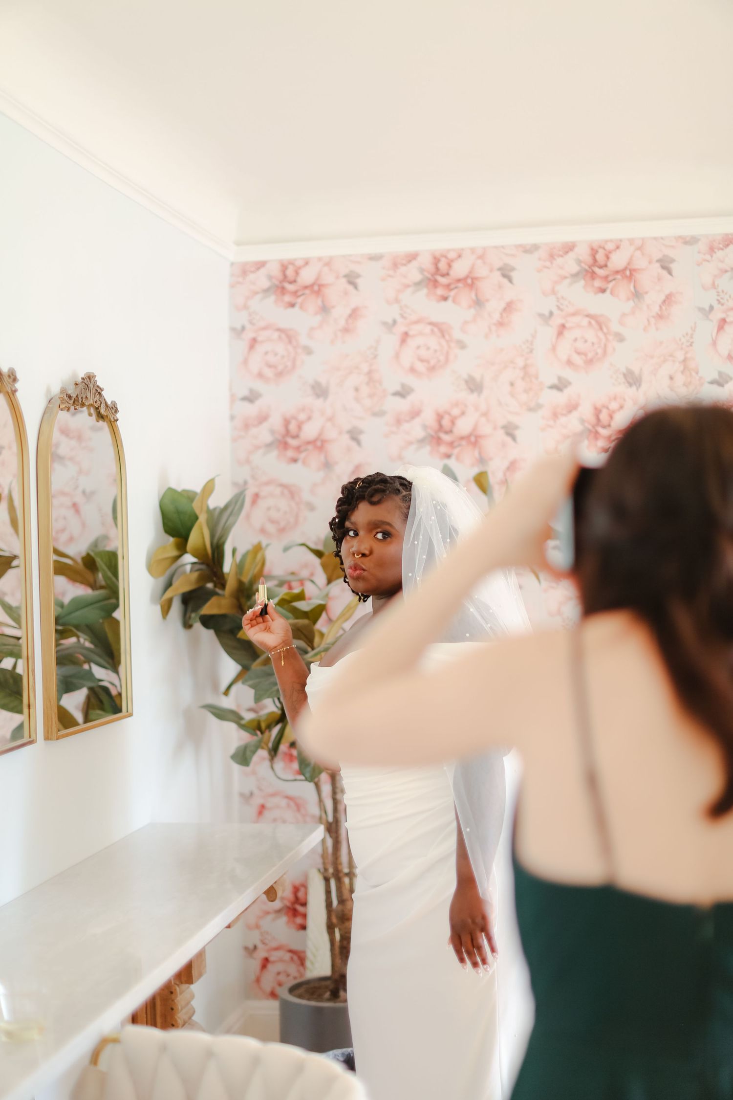 A bride in an elegant white dress and veil stands in a room with floral wallpaper and a decorative mirror.