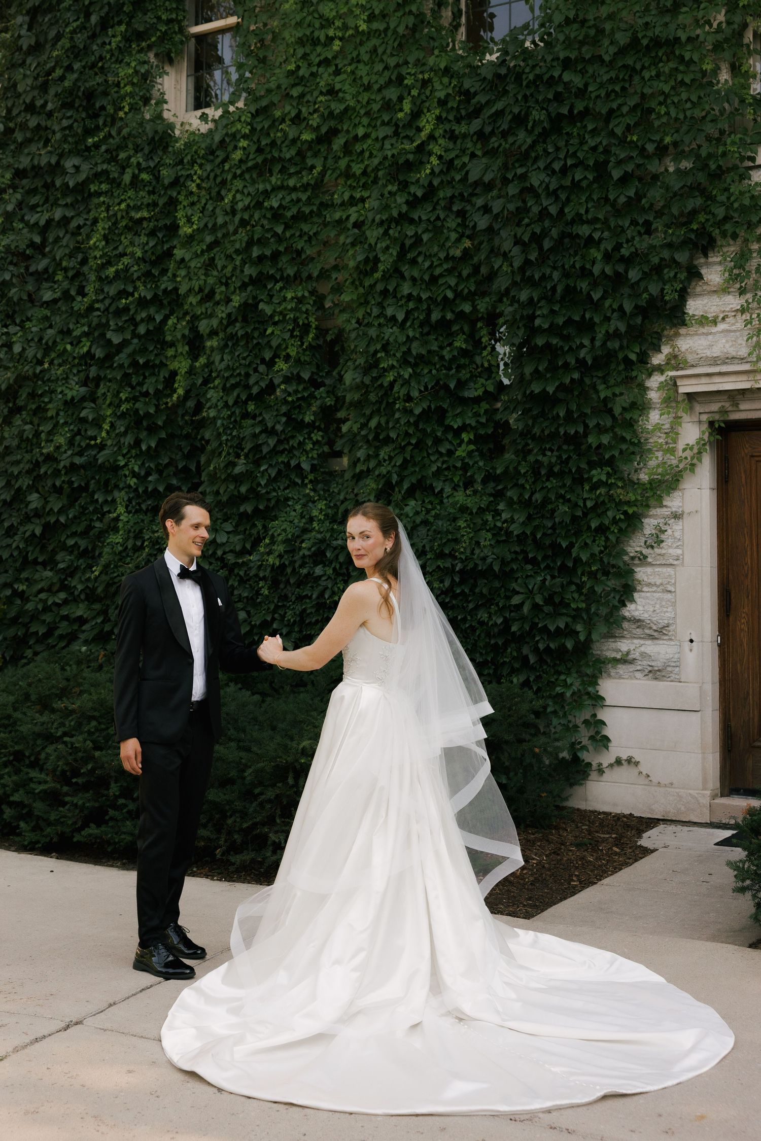 A bride in a white gown and veil holds hands with a groom in a black tuxedo before an ivy-covered stone wall.