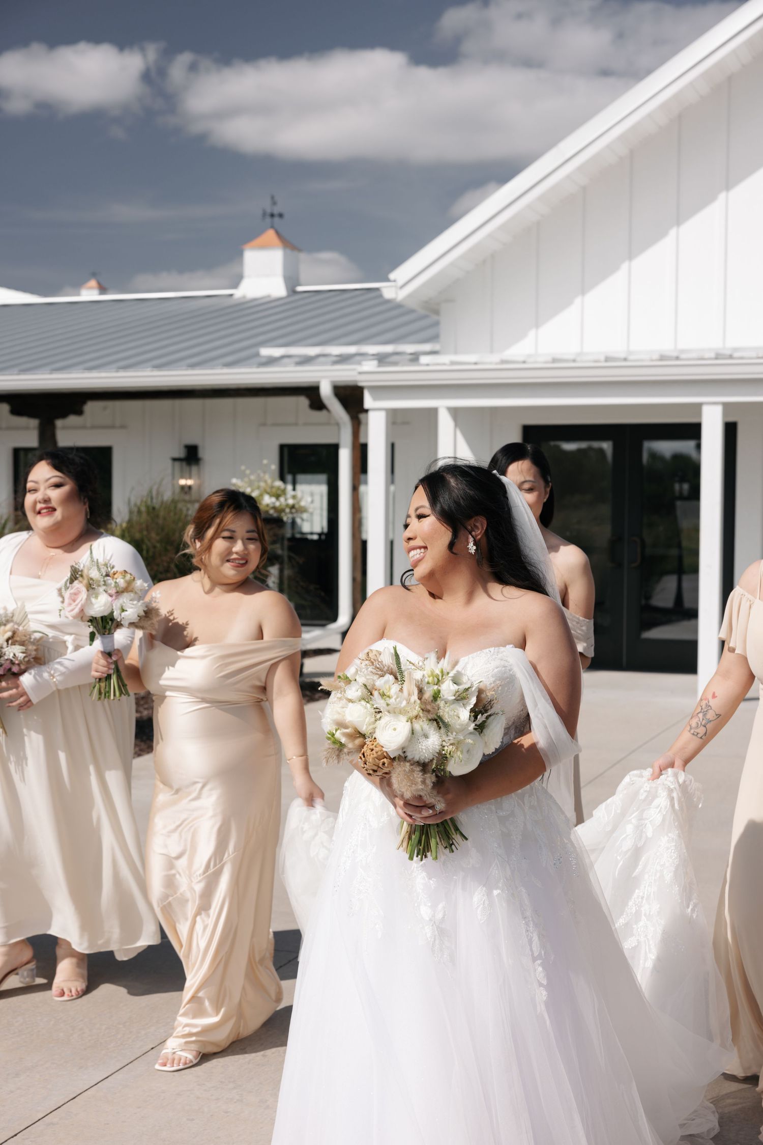 A bridal party in white and champagne colored dresses laughs and poses together on a white porch.