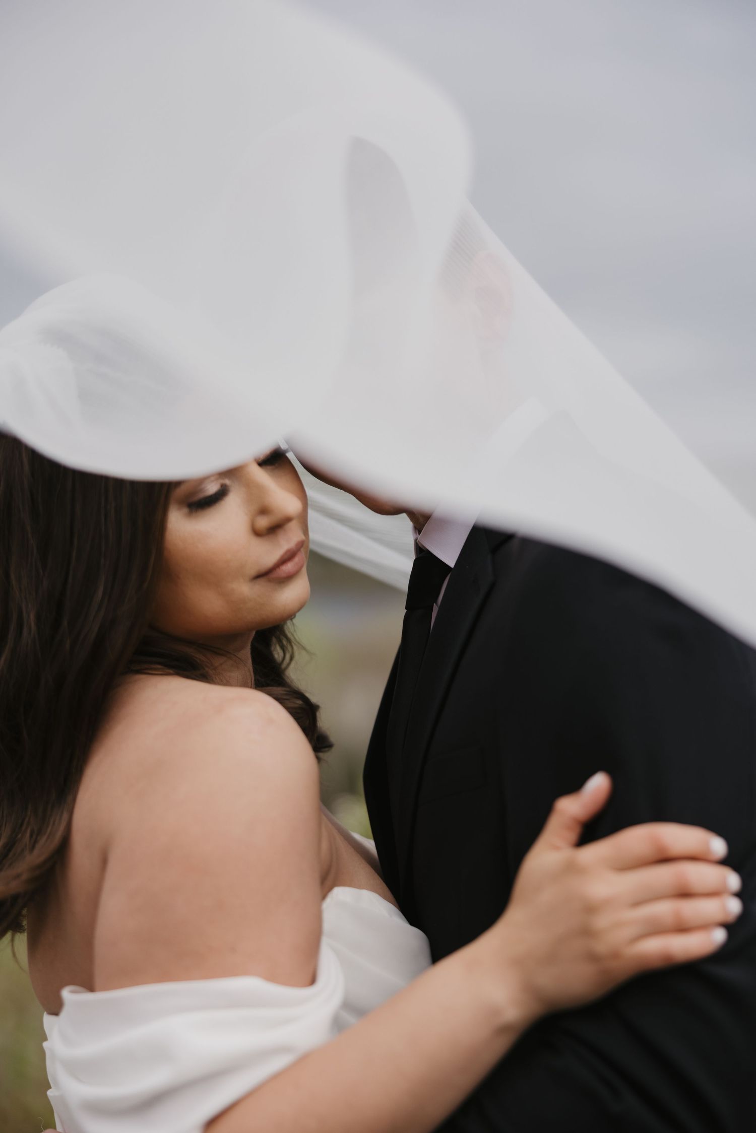A romantic wedding moment captured as a white veil flows over an intimate embrace between newlyweds.