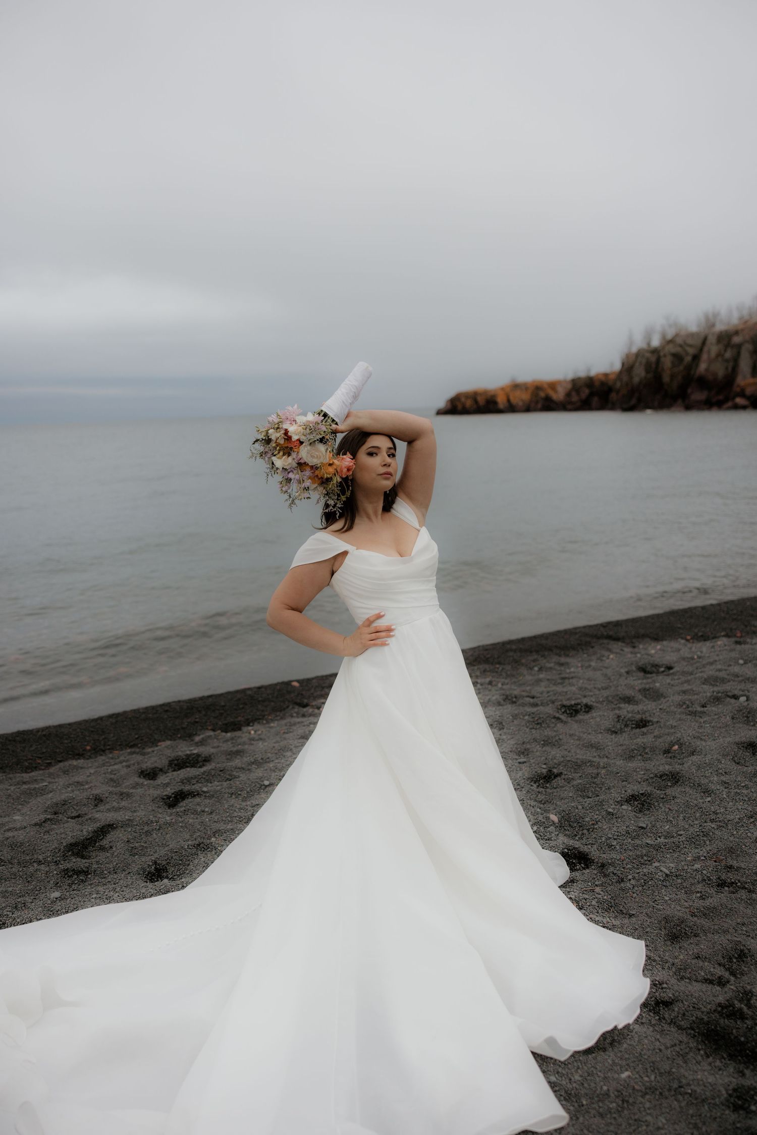 A wedding dress photoshoot on a moody black sand beach with dramatic cloudy skies and coastal cliffs in the background.