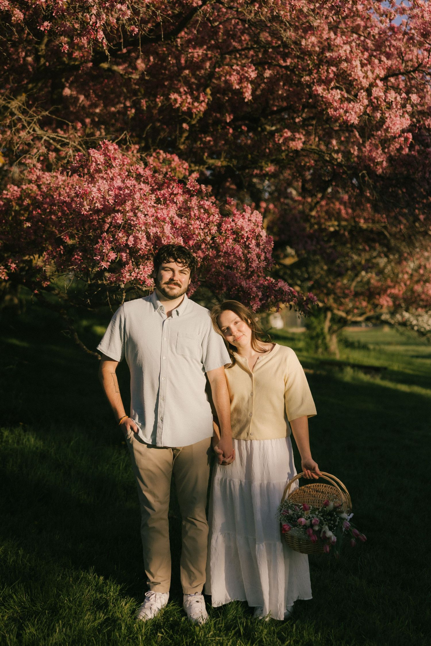 Couple standing together under blooming pink cherry blossom tree, woman holding flower basket, golden hour light.