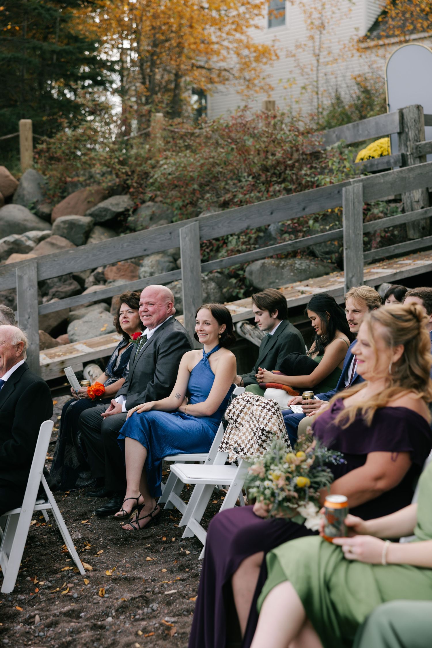 Wedding guests seated on white chairs during an outdoor autumn ceremony with fall foliage in the background.