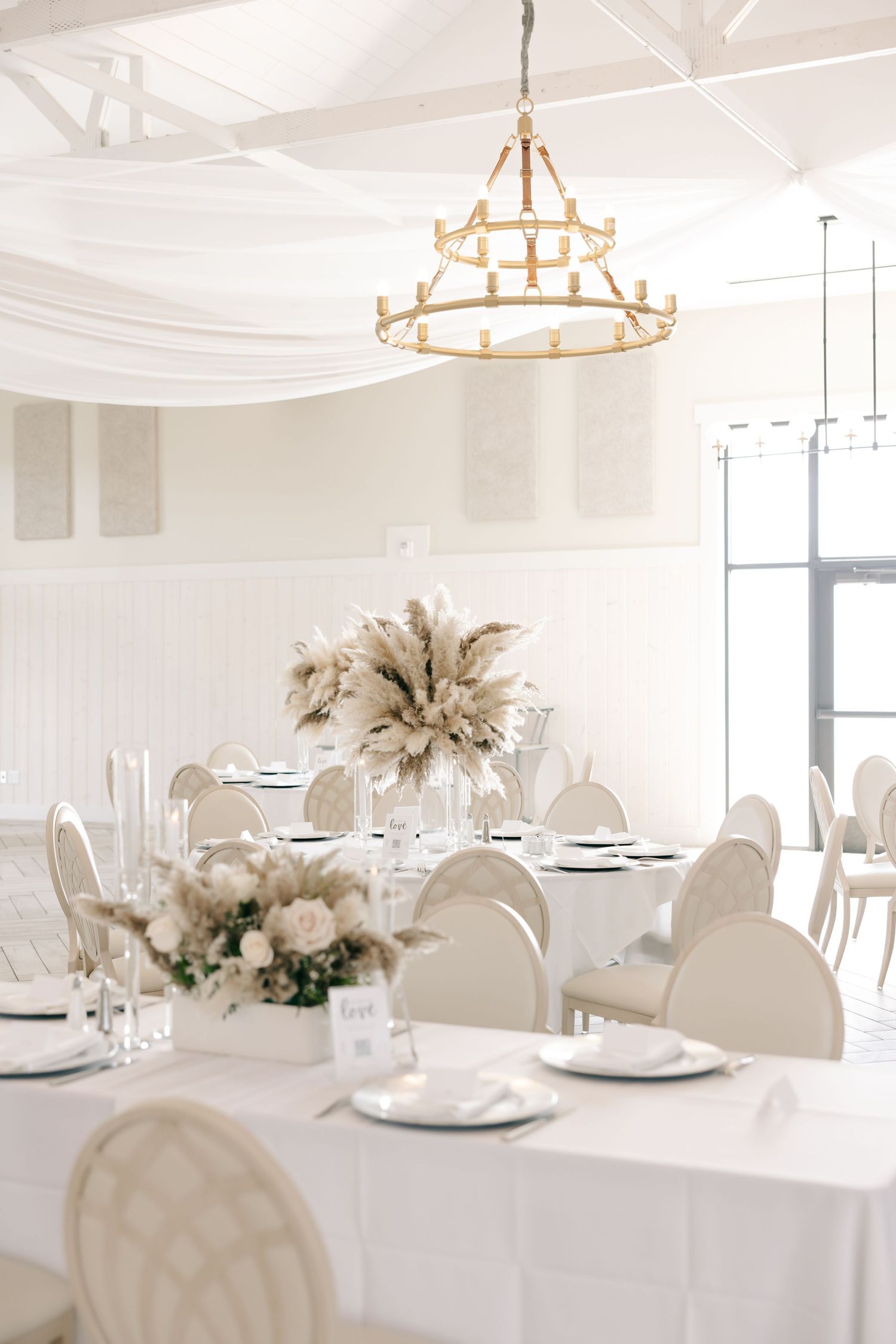 Elegant white wedding reception venue with round tables, white chairs, and rustic chandeliers creating a bright airy atmosphere.