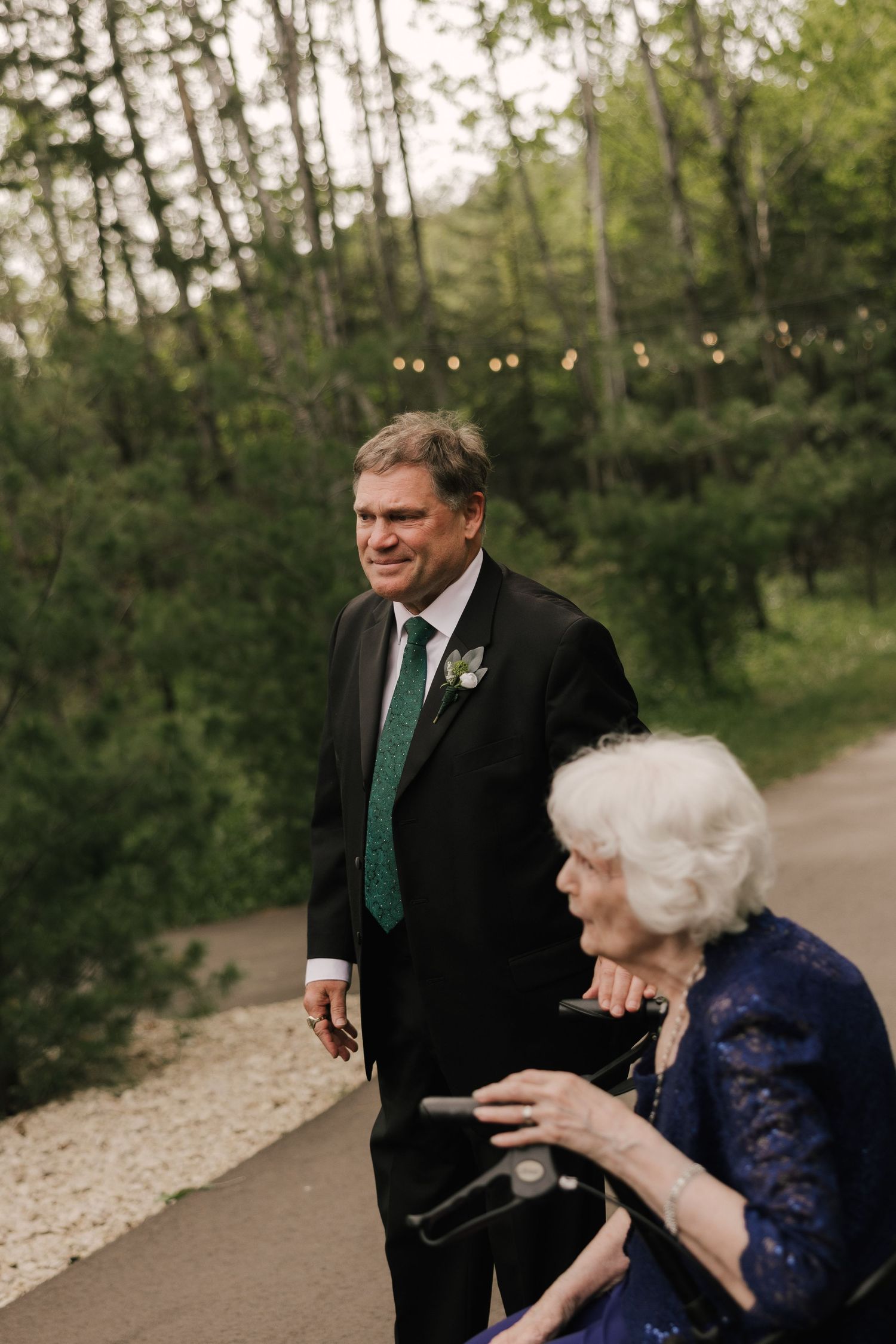 A man in a black suit with green tie and boutonniere stands beside an elderly woman in a blue lace dress outdoors.