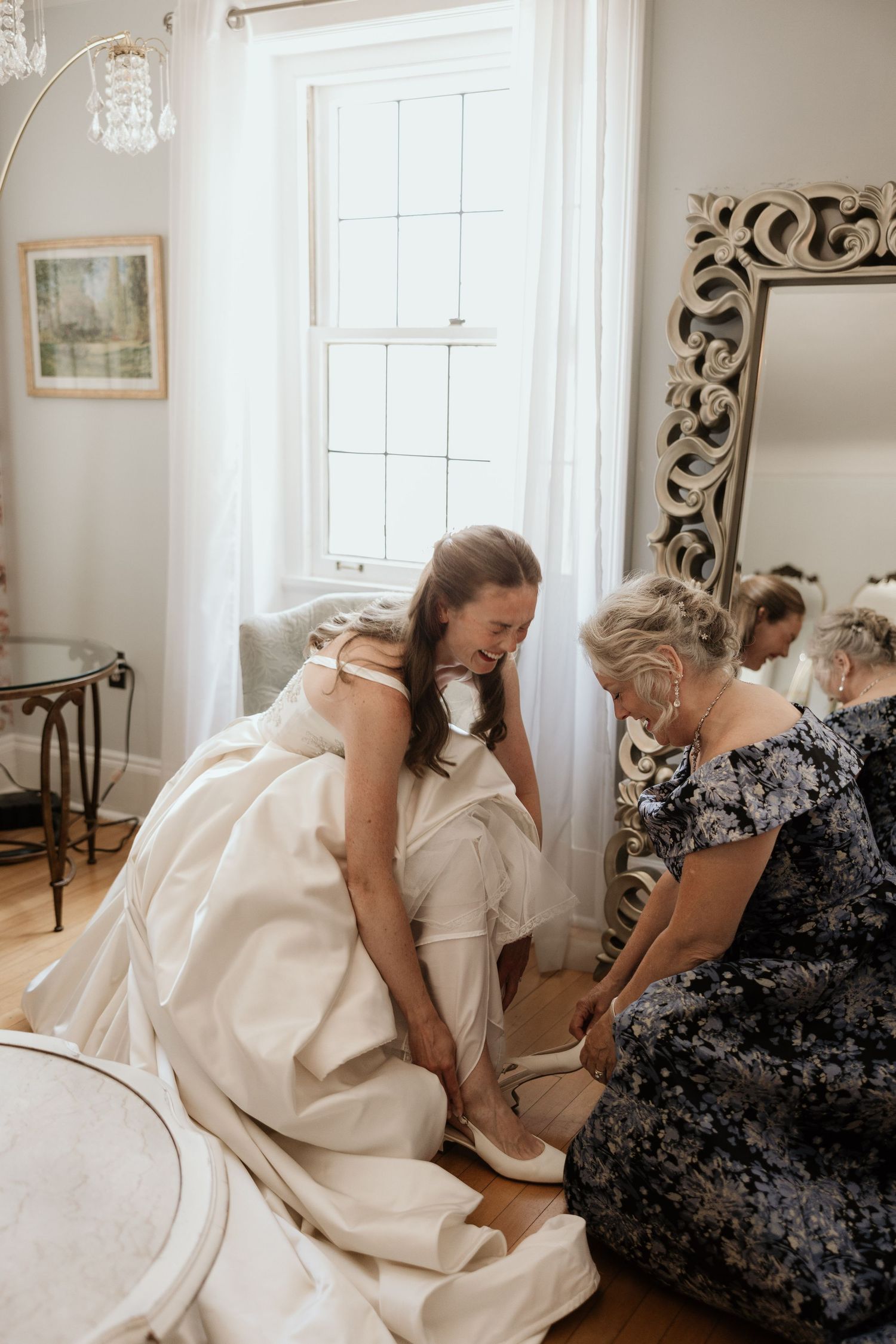 Bride in white gown getting shoes put on by older woman near ornate mirror in bright elegant room.