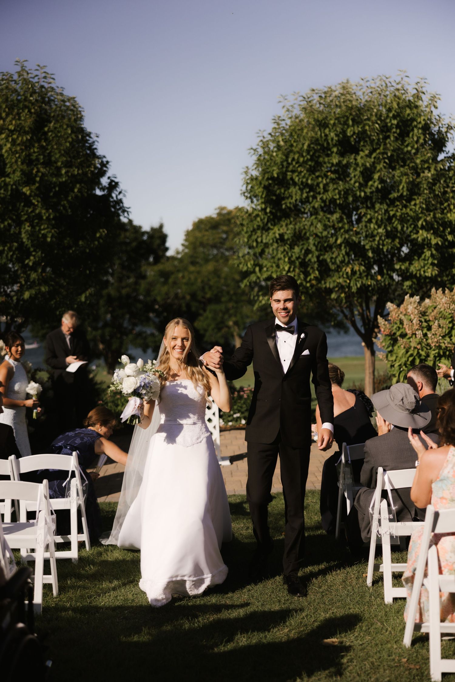Newlywed couple walks down outdoor wedding aisle, bride in white gown holding bouquet, groom in black tuxedo, guests seated on white chairs.