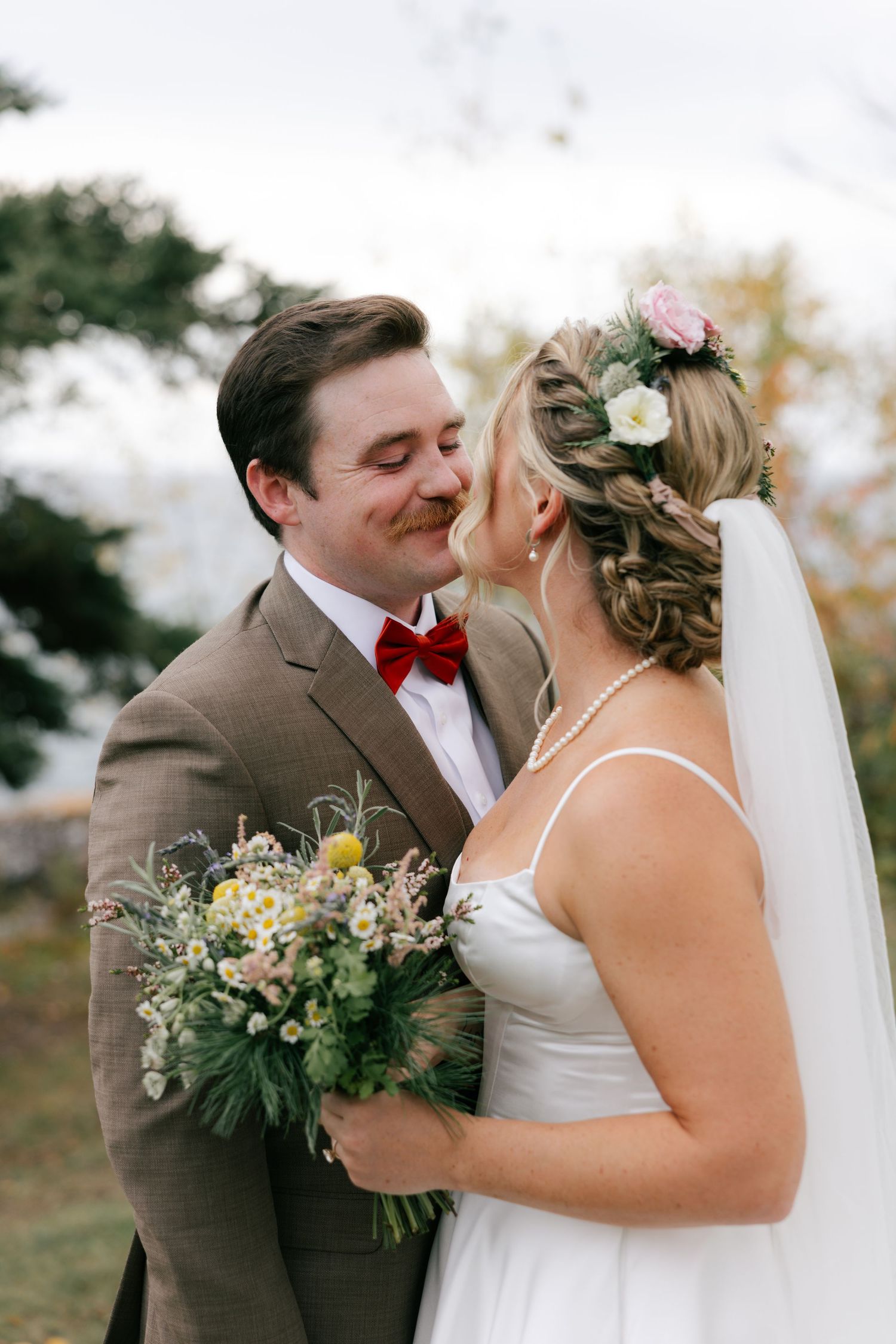 A romantic outdoor wedding scene with a couple sharing tender kisses while holding a rustic bouquet of greenery and flowers.