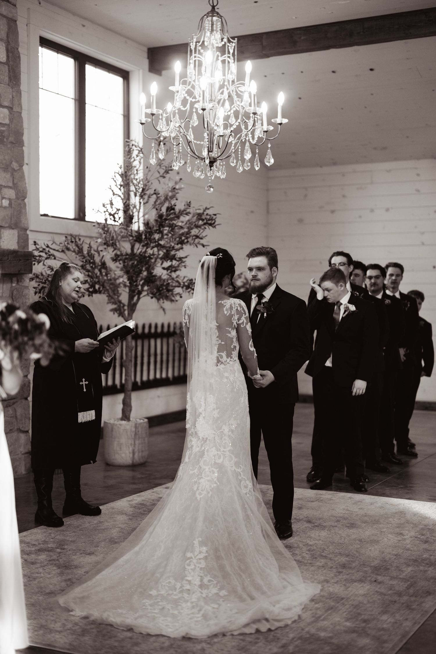A wedding ceremony in a rustic white venue with elegant chandeliers and a line of formally dressed guests watching.