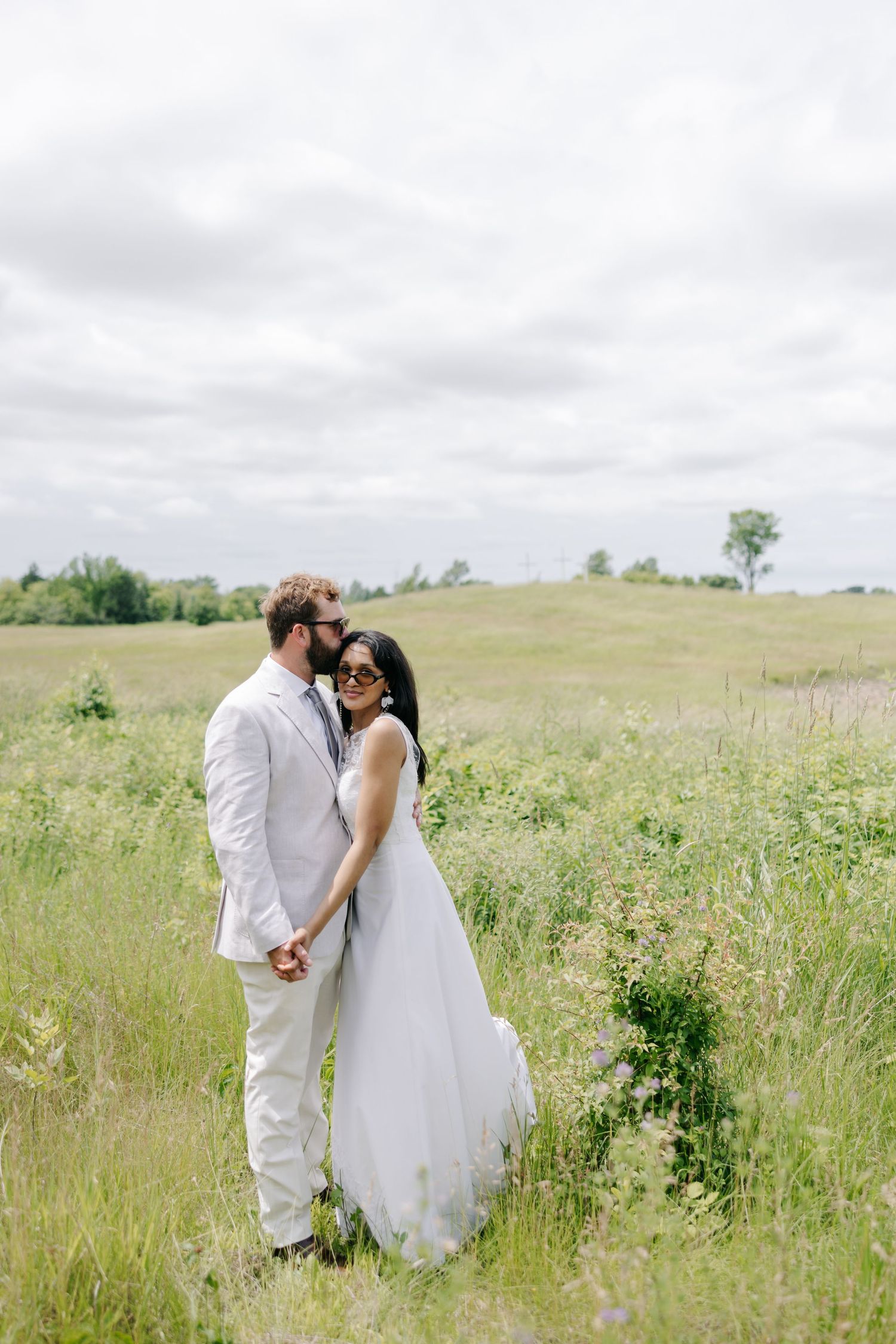 A romantic couple in wedding attire embrace in a serene meadow with wildflowers under an overcast sky.