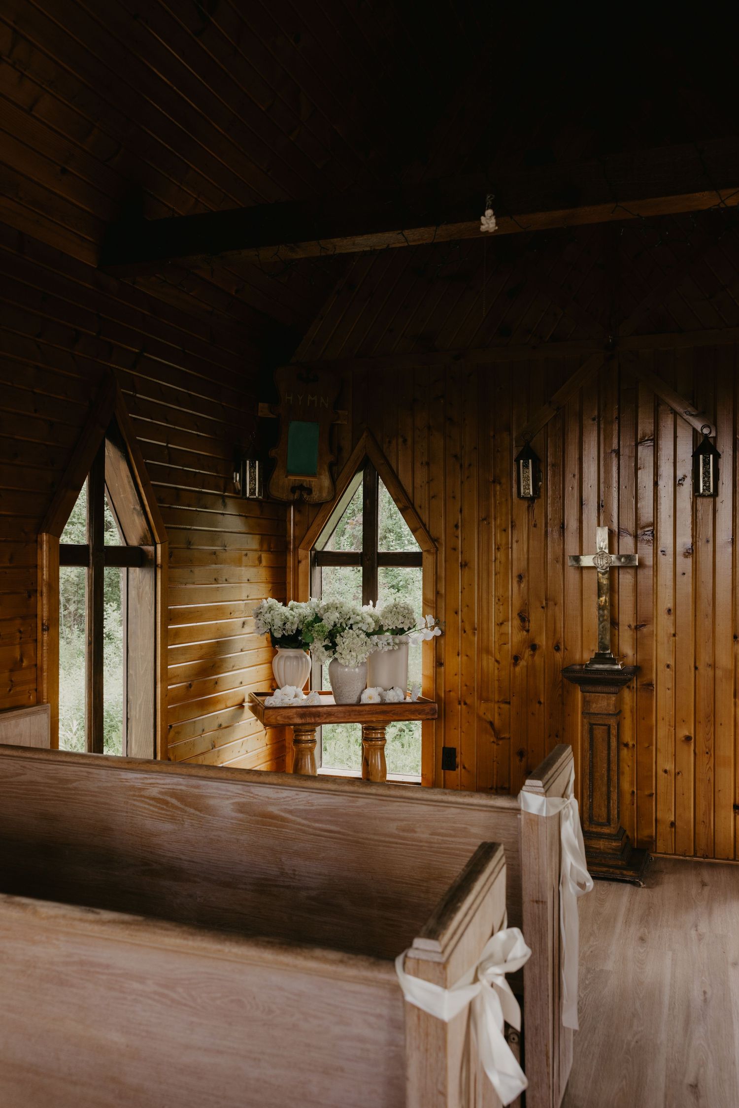 Interior view of a rustic wooden chapel with paneled walls, hanging chandeliers, and white pews in warm natural light.