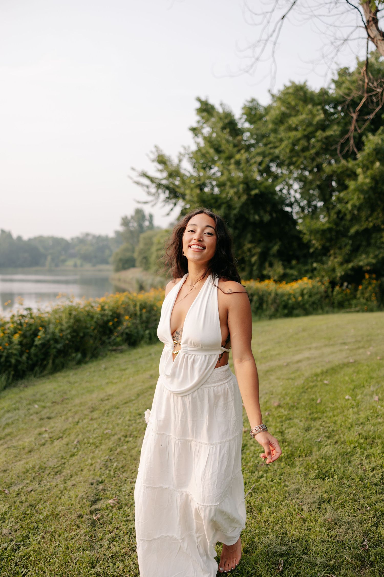 A woman in a flowing white maxi dress walks along a grassy hill near a lake on a sunny summer day.