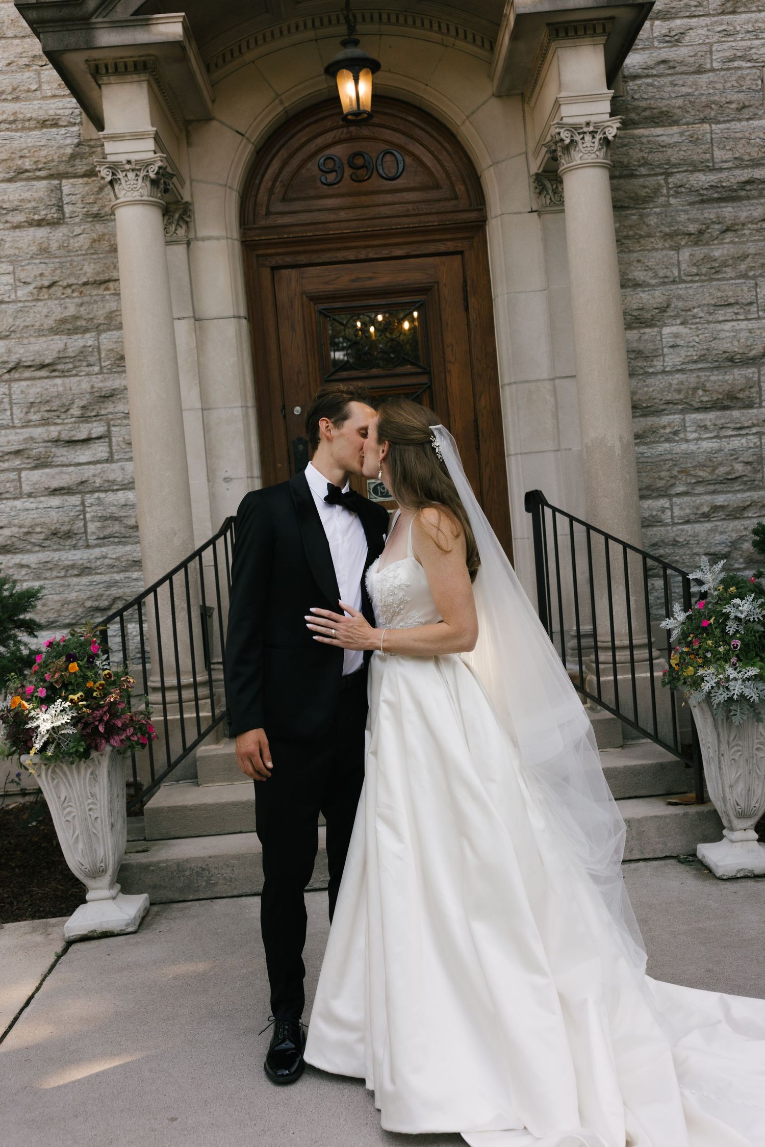 Bride and groom kissing on steps of elegant stone building, bride in white ballgown with veil, groom in black tuxedo.