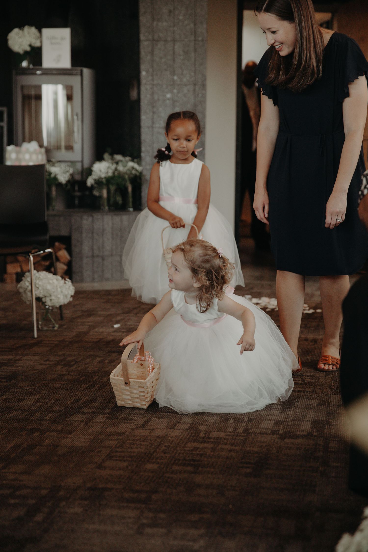 Two flower girls in white dresses play with basket during wedding ceremony.