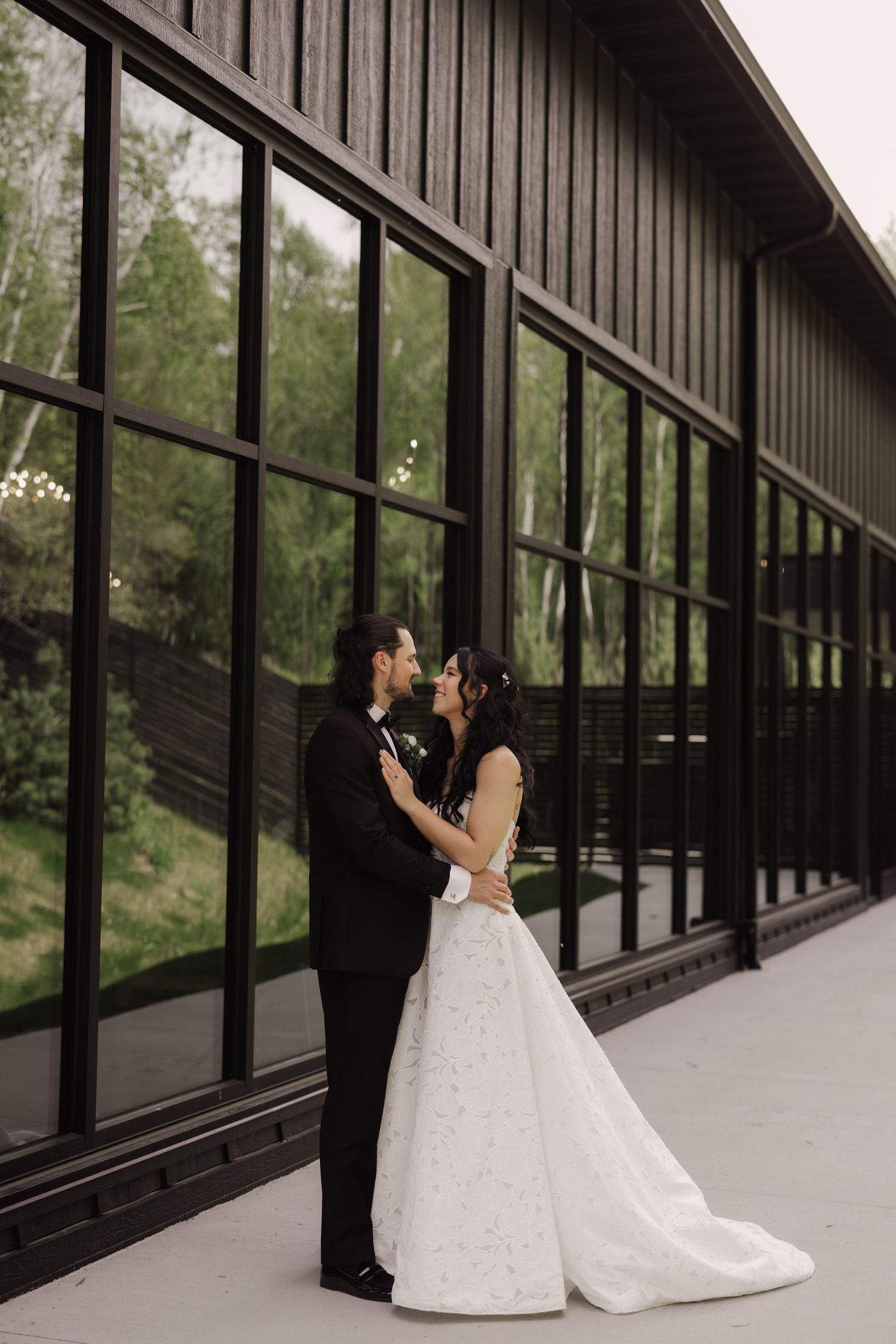 Bride and groom share a romantic kiss outside a modern black-framed glass building surrounded by greenery.