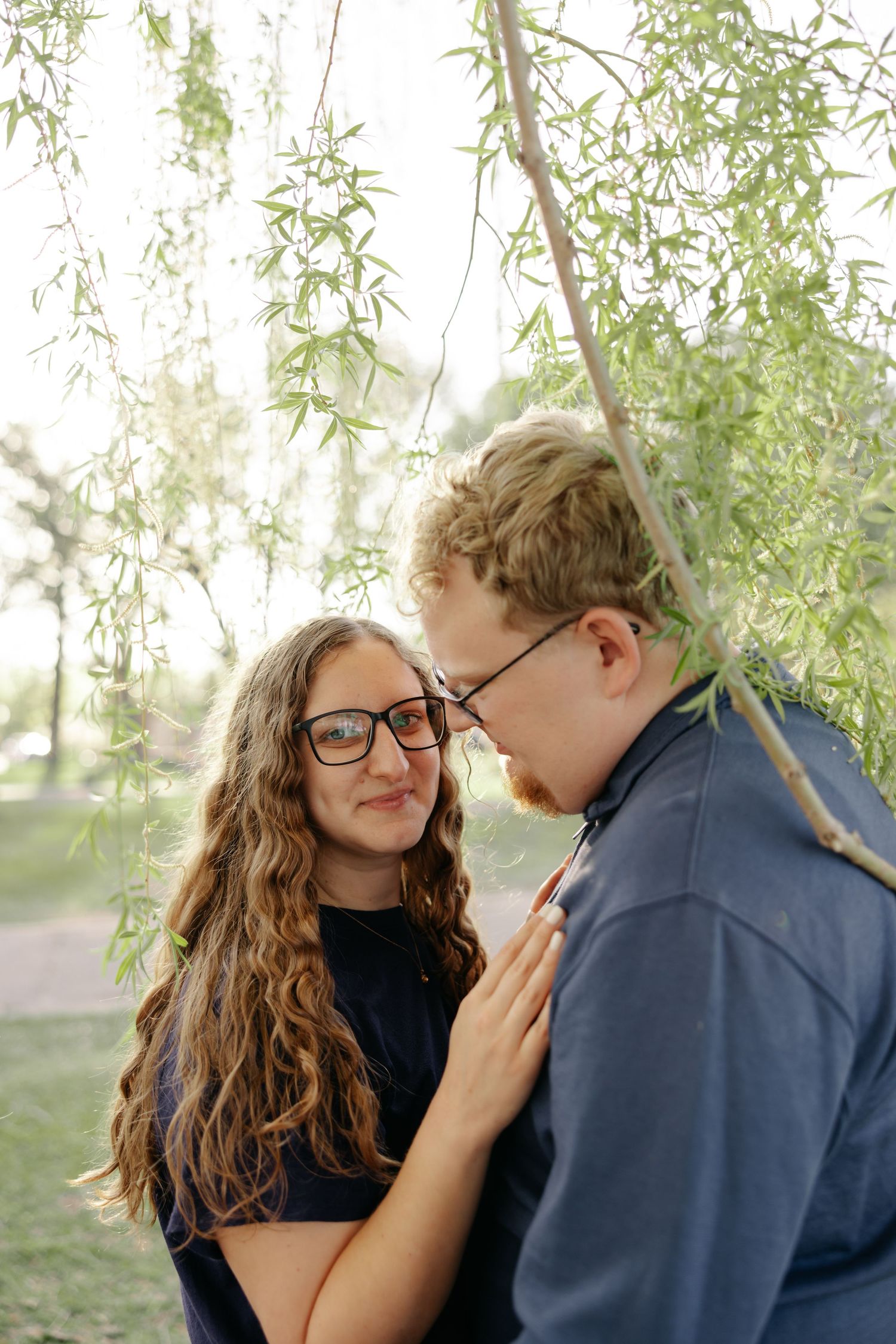 A couple shares an intimate moment beneath drooping willow tree branches in a romantic outdoor setting.