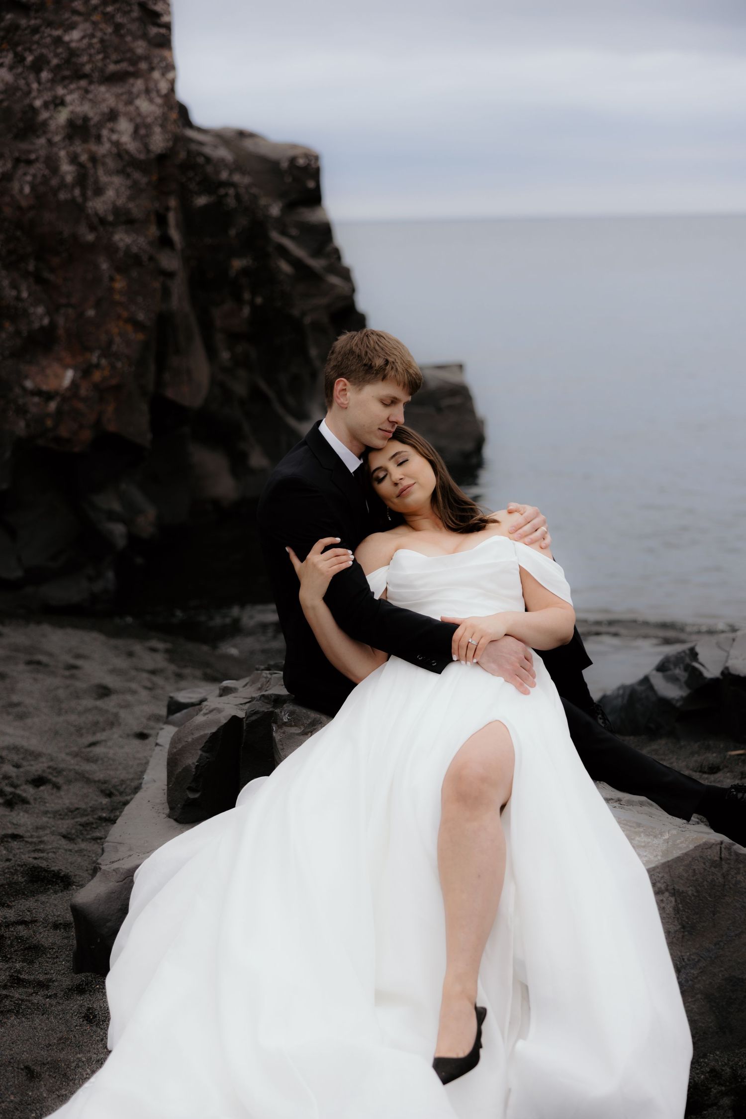 A romantic wedding couple embrace on dark rocky cliffs overlooking a misty ocean.