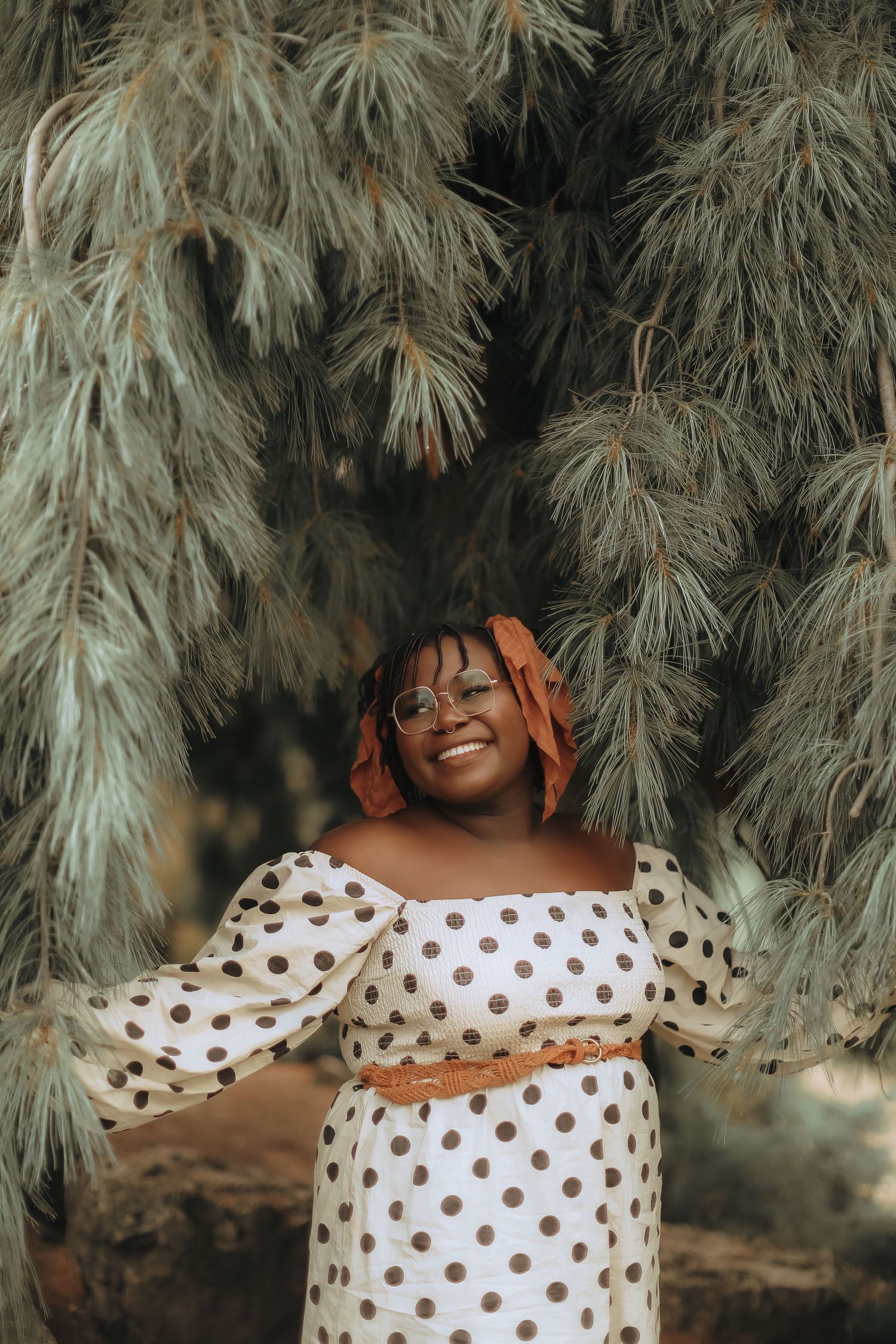 A joyful person in a polka dot dress poses beneath drooping willow branches.