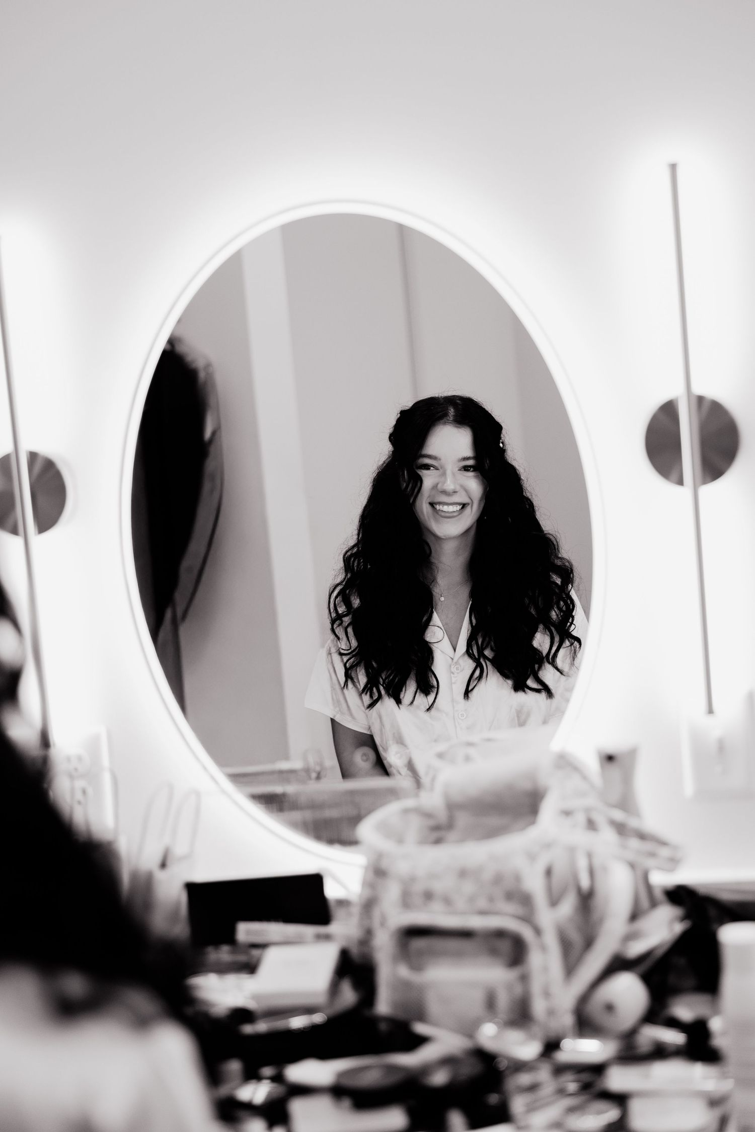 Black and white photo of smiling bride with long wavy hair reflected in oval vanity mirror.