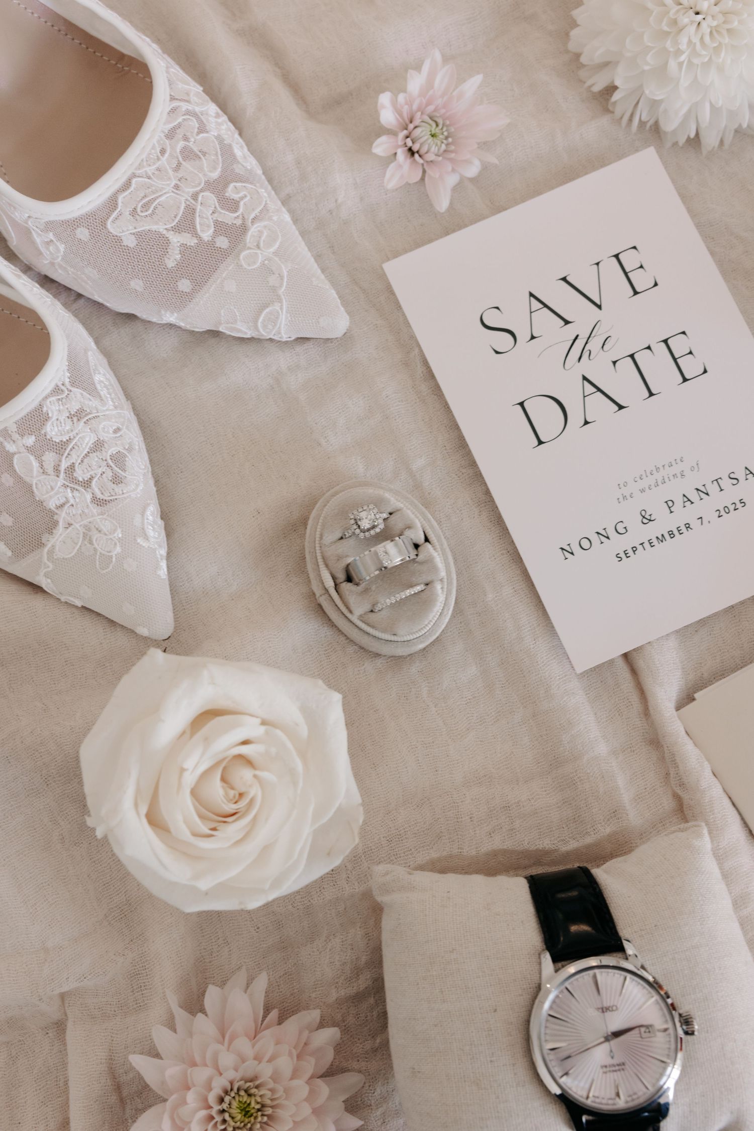 Elegant wedding details including lace shoes, save the date card, rings, white rose, and wristwatch on beige fabric.
