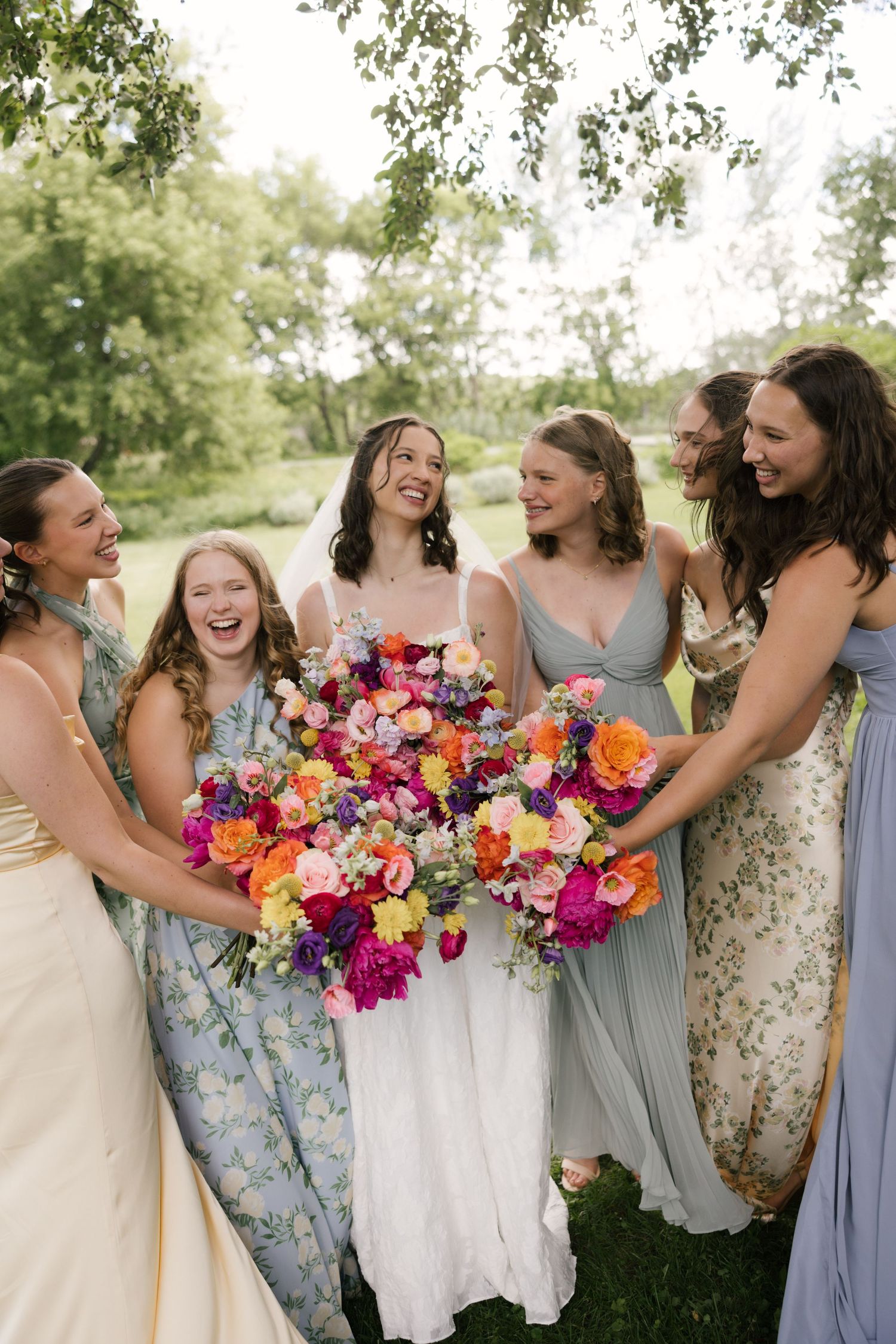 Bride in white gown holds vibrant colorful bouquet surrounded by laughing bridesmaids in mismatched dresses outdoors.