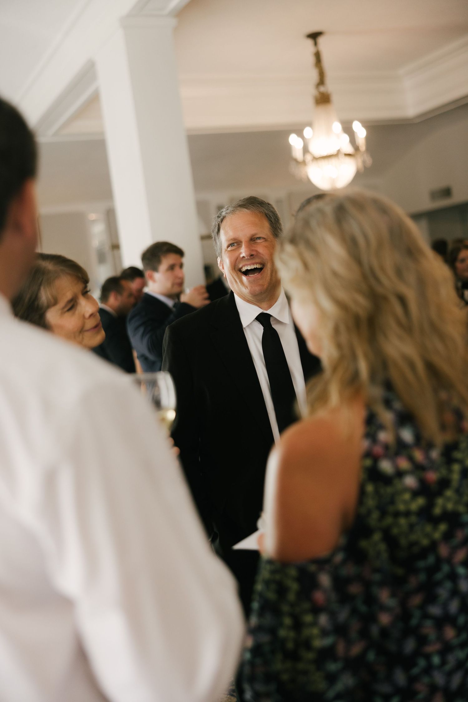 Man in black suit laughing joyfully at an elegant indoor social gathering with chandelier lighting.