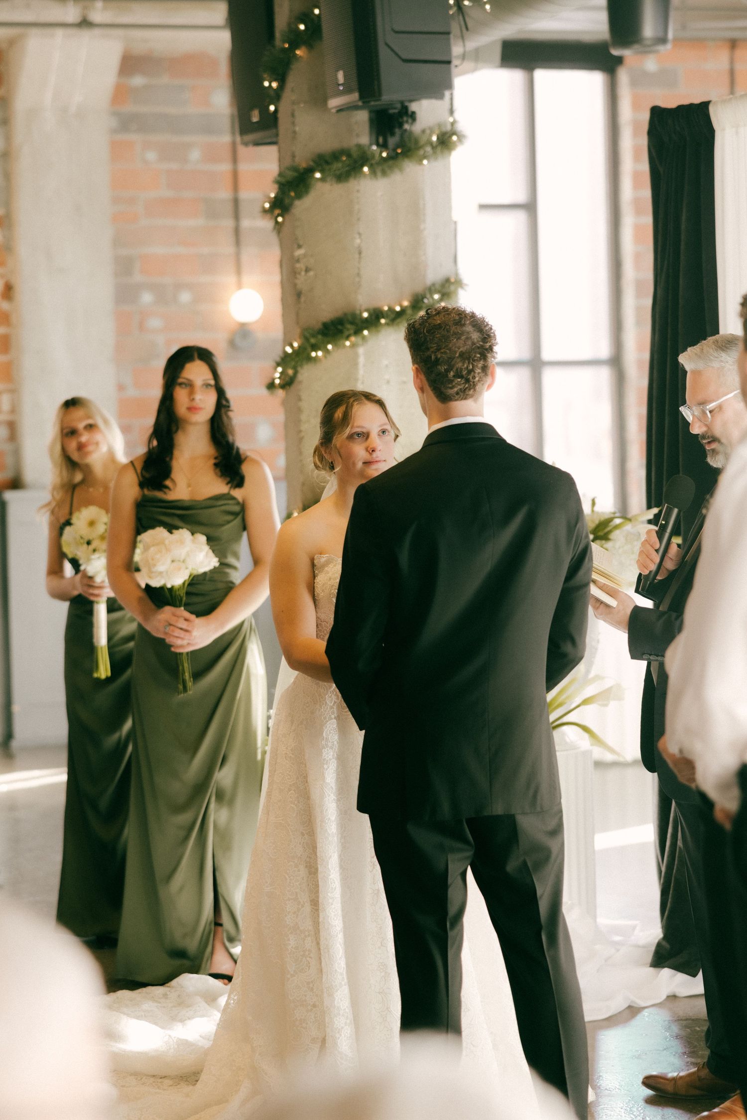 Wedding party members in black and olive formal attire dance and pose in a bright industrial venue with holiday decor.